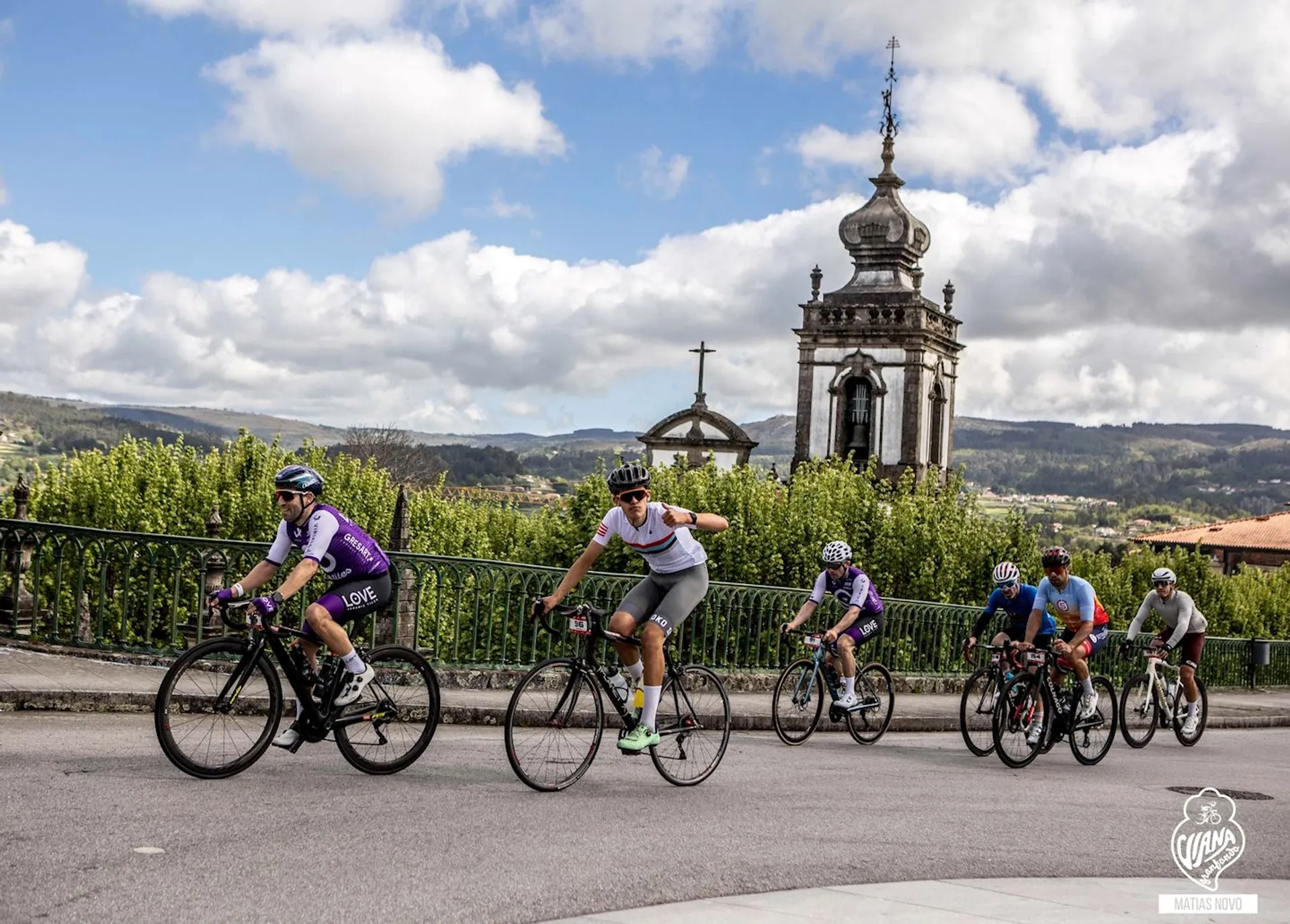 In the image, there is a group of cyclists riding on a road. In the background, there is an old-style building with a prominent bell tower featuring a dome and a cross at the top, which suggests it might be a church or a cathedral. The landscape behind the cyclists is scenic with lush greenery and trees, and it appears to be a countryside or rural area. The sky is partly cloudy, indicating a day with mixed weather. All the cyclists are wearing cycling gear and helmets, indicating they are engaged in a sporting or recreational activity, possibly a group ride or cycling event.