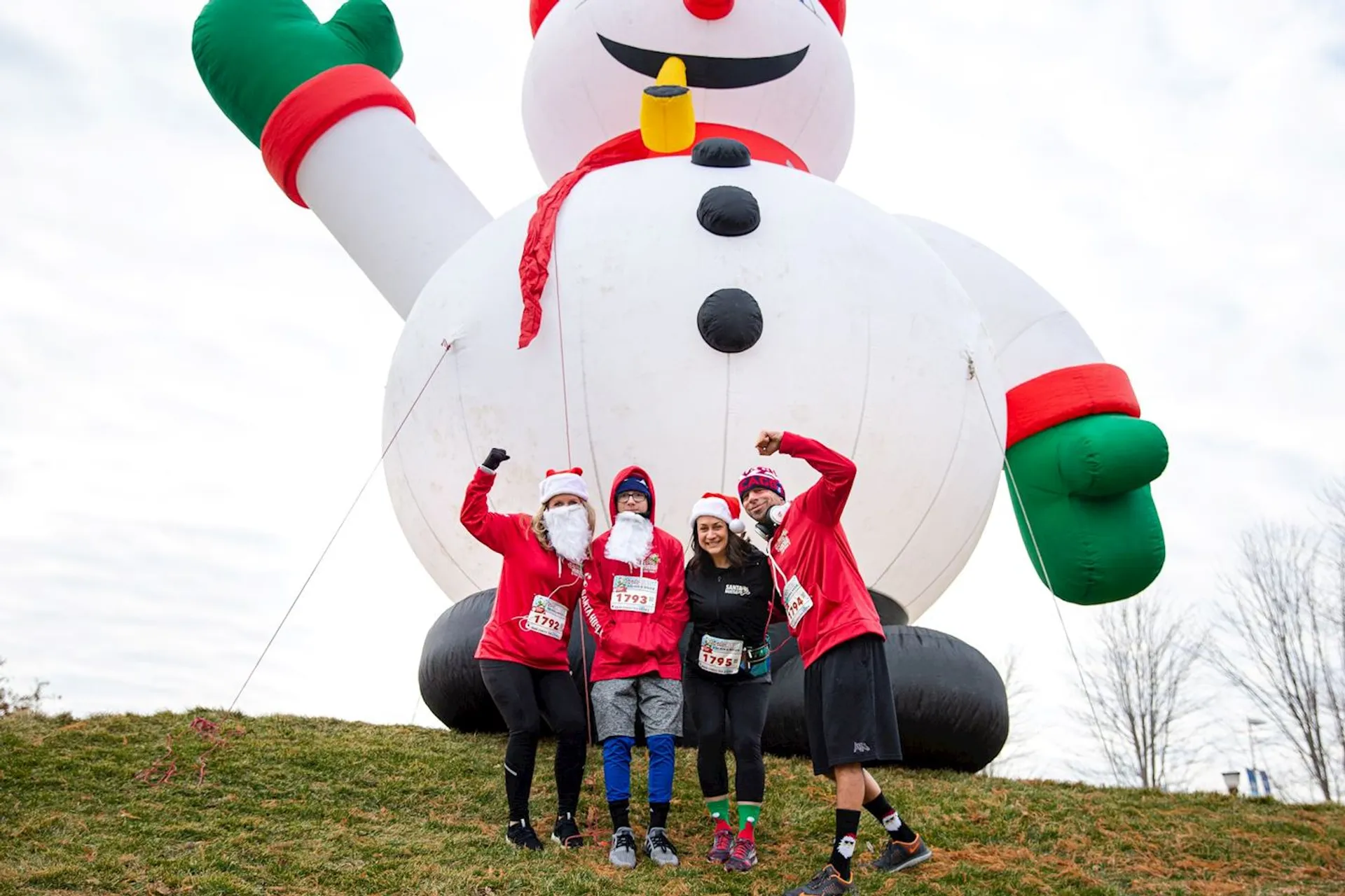 The image shows four people posing in front of a large inflatable snowman. The individuals are wearing festive attire, such as Santa hats and beards, and are likely participating in a holiday-themed event. They appear to be on a grassy area.