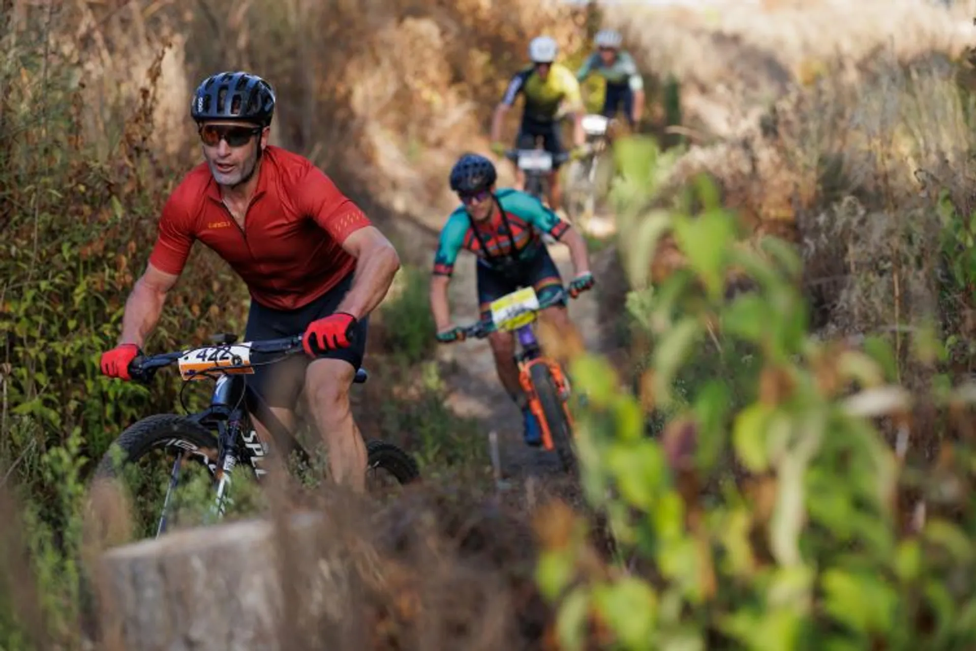 The image shows several people riding mountain bikes on a narrow, dirt trail surrounded by vegetation. The riders are wearing helmets and cycling gear. The environment suggests an outdoor, off-road cycling activity.