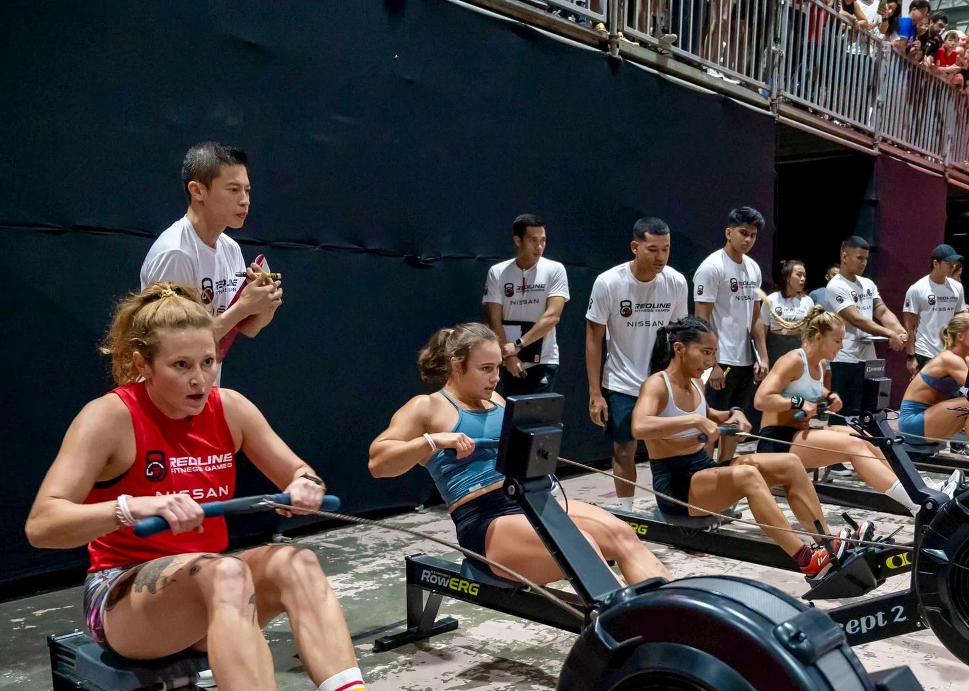 The image shows a group of athletes participating in a rowing competition. They are using rowing machines, lined up in a row, with several individuals standing nearby, possibly coaches or judges. The setting appears to be an indoor sports venue.