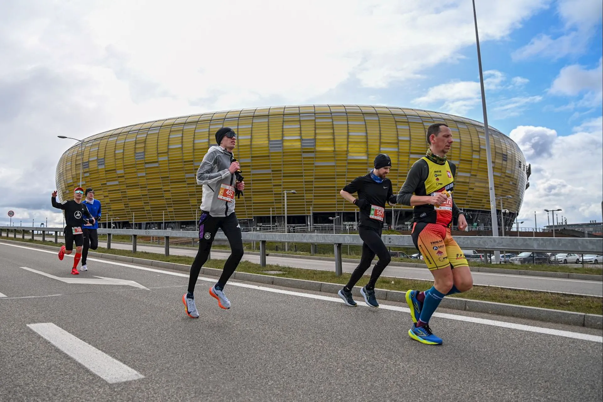 The image shows a group of people running on a road, likely participating in a marathon or race event. They are wearing athletic clothing and race bibs. In the background, there is a large, yellow-gold stadium. The sky is partly cloudy.