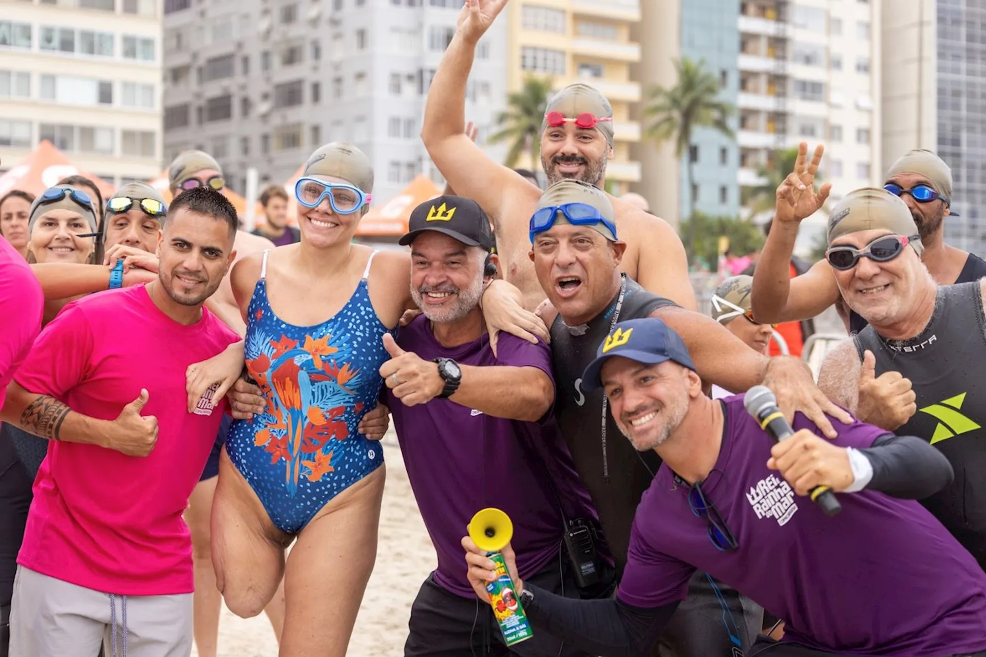 This image shows a group of people on a beach, some wearing swimming gear and swim caps. They appear to be participating in a water-related event or activity, like an open water swim. A few people are holding a horn and wearing event t-shirts, suggesting they might be organizers or participants in this event. They all seem to be in good spirits, smiling and posing for the photo.