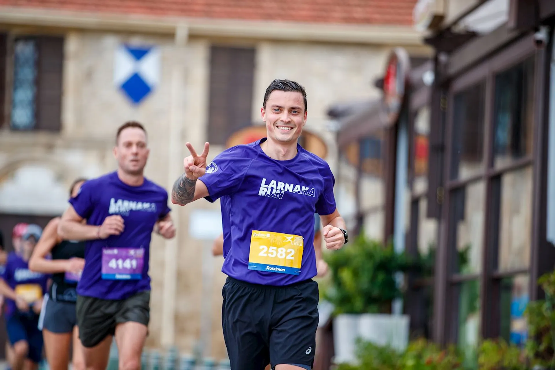 The image shows several people participating in a running event. One person in the foreground is making a peace sign and wearing a purple shirt with "Larnaka Run" on it, along with a numbered bib. Other runners can be seen in the background, and the setting appears to be an outdoor area.