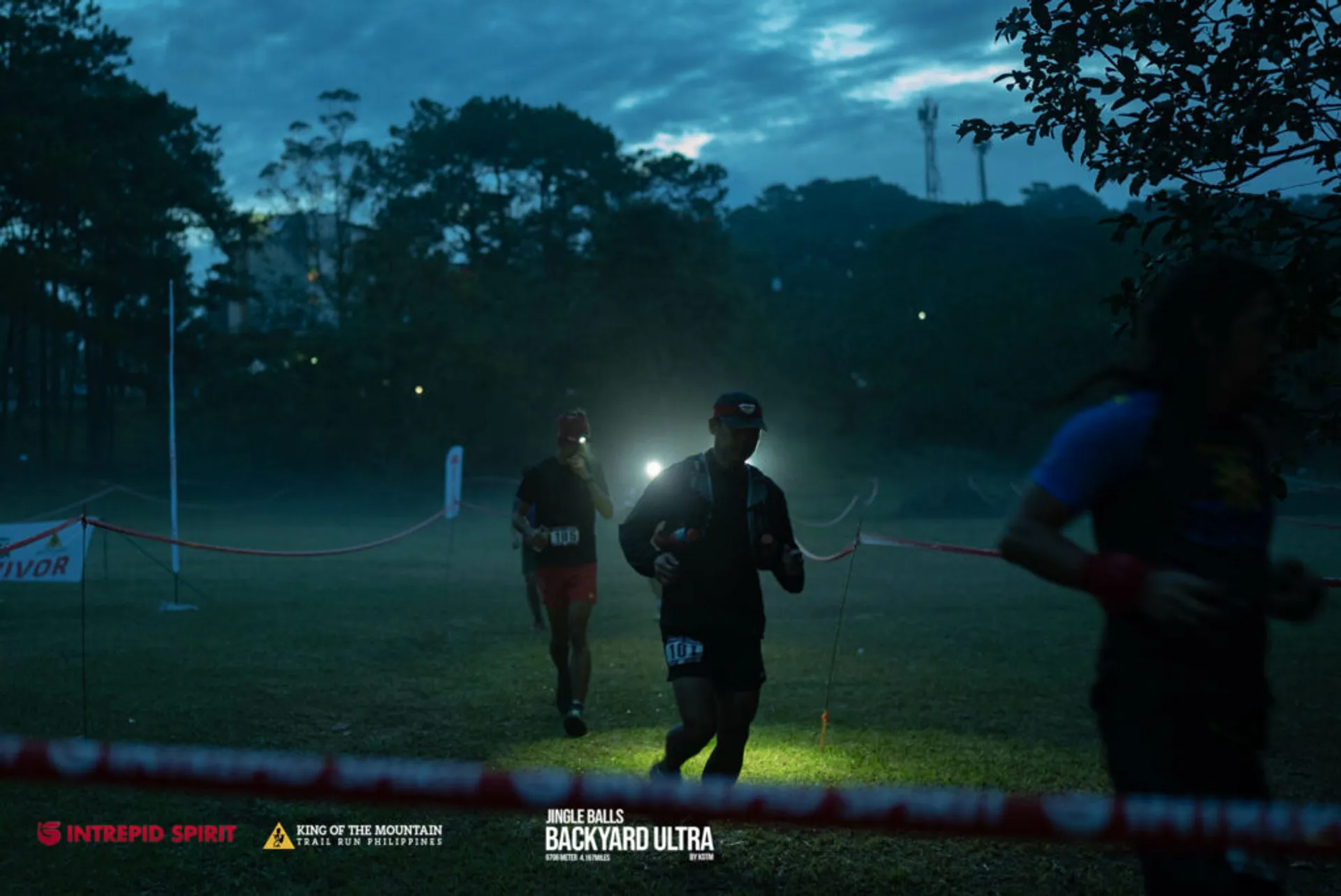 The image depicts a night-time running event, possibly an ultra marathon or endurance race. There are runners equipped with headlamps to illuminate their path, as visibility is low. The scene indicates an outdoor, grassy area surrounded by trees, with the early morning or evening light creating a dim ambiance. Banners and tape mark the course, ensuring runners adhere to the designated path. Visible emblems and sponsors such as "Survivor," "INTREPID SPIRIT," "KING OF THE MOUNTAIN SERIES," and "INEAGLES BACKYARD ULTRA" suggest the official nature of the race. The participants appear focused and determined, indicating the competitive and challenging nature of the event.