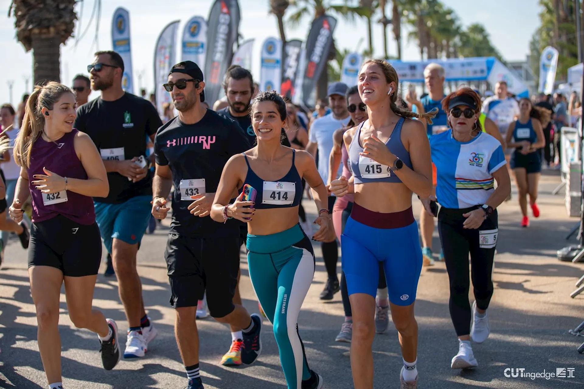 The image shows a group of people participating in a running event. They are wearing athletic clothing and bib numbers, indicating they are part of a race or marathon. The runners seem to be in good spirits, with some of them smiling and appearing to enjoy the competition. The setting is outdoors, with palm trees in the background, suggesting a warm climate. Banners and flags for what appears to be sponsors or organizers of the event line the running path, creating a lively atmosphere. The focus and energy of the participants suggest a community or sporting event centered on fitness and wellbeing.