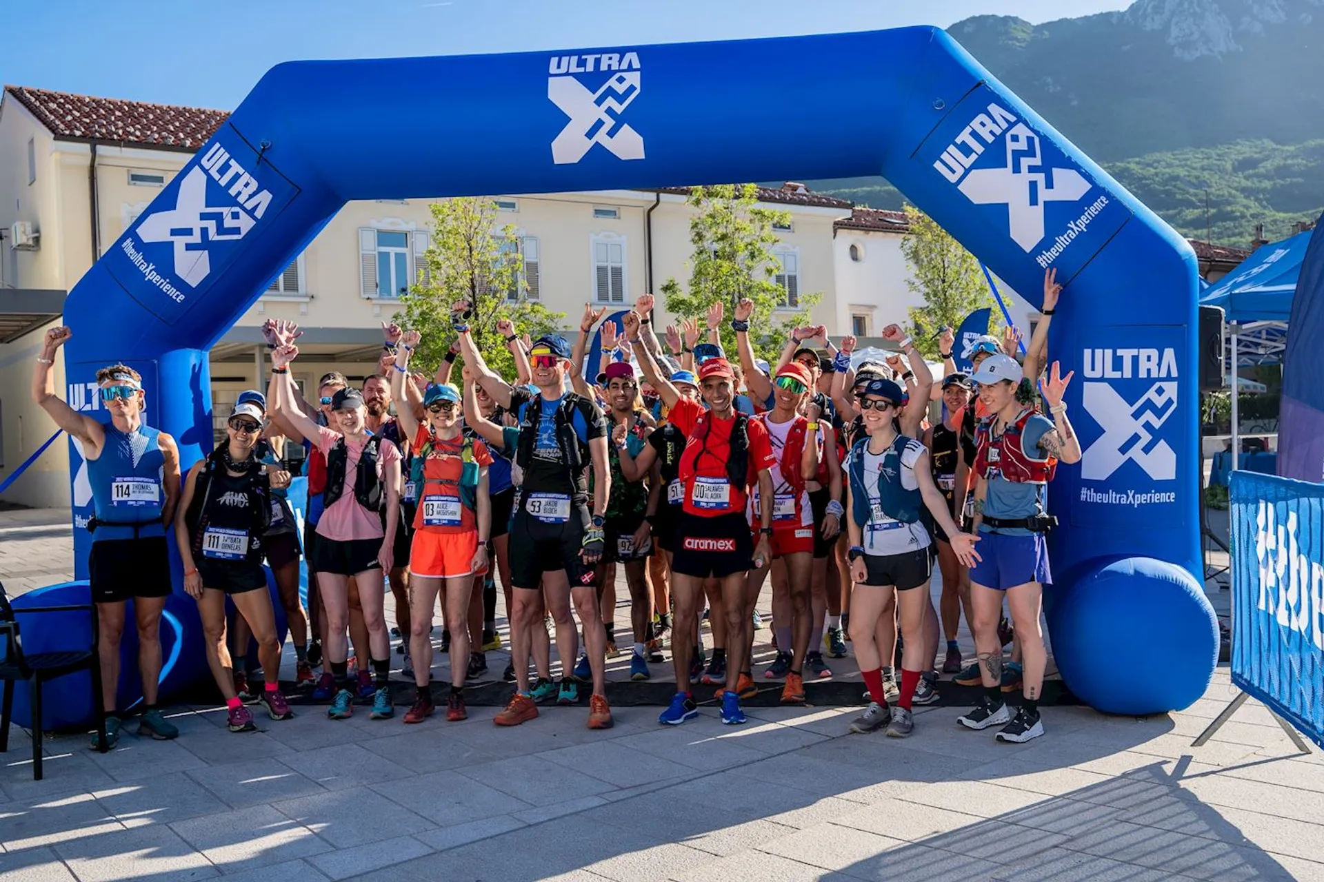 The image shows a group of happy and enthusiastic runners gathered at the starting line of an outdoor running event, specifically an ultra-marathon race as indicated by the "ULTRA" branding on the inflatable arch. They are wearing athletic clothing, hydration packs, race bibs, and various running gear suitable for long-distance events. The runners are standing in a scenic location with a clear sky and a mountainous backdrop, suggesting that the race is likely to take place in a location with natural trails or paths. They have their arms raised, some making celebratory gestures, indicating excitement and readiness for the race ahead.