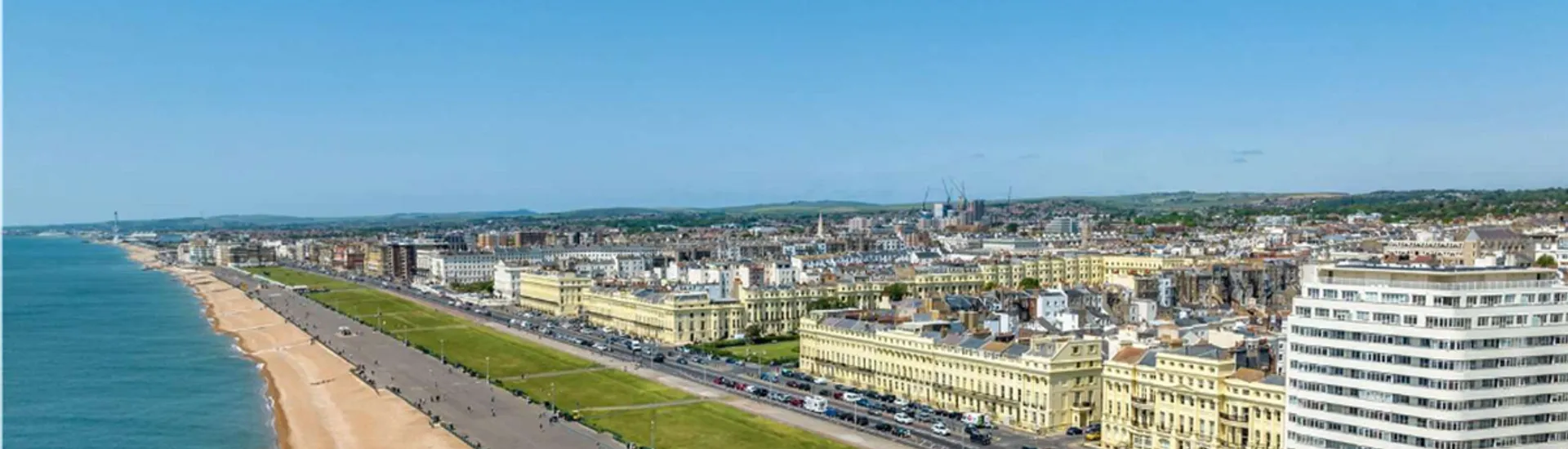 The image shows a panoramic view of a coastal city. You can see a long beach with what appears to be a promenade lined with buildings. The architecture suggests a mix of residential and commercial properties, possibly from the Victorian or Georgian era, which is typical in certain British seaside towns. The overall layout, with the sea on one side and the city expanding into the distance, contributes to a scenic urban landscape. The weather is clear, offering a blue sky and calm sea, which enhances the attractiveness of the place.