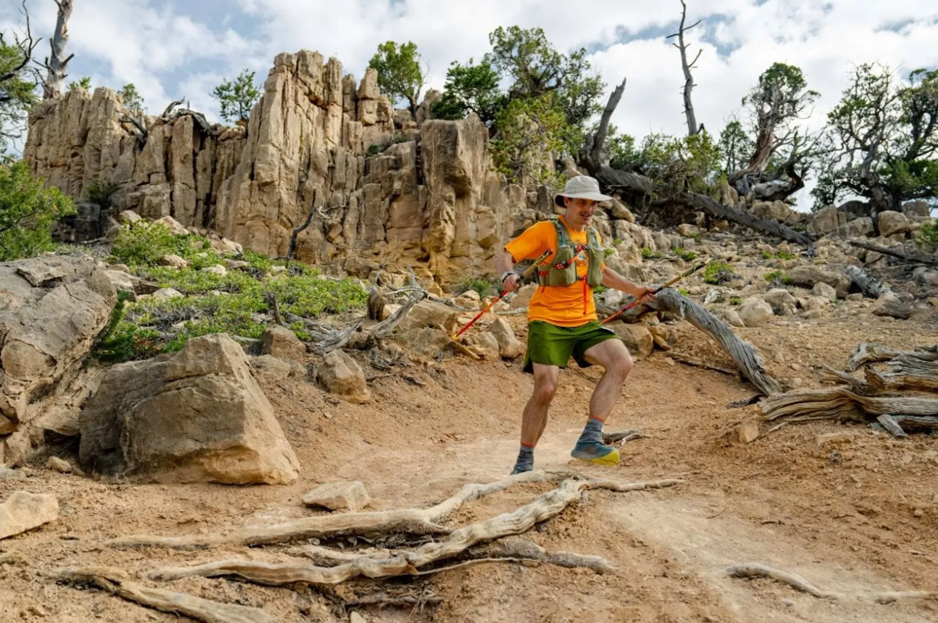 The image shows a person hiking on a rocky trail. They are wearing an orange shirt, green shorts, a hat, and carrying walking poles. The landscape features rugged terrain with rocks and trees, and the sky is partly cloudy.