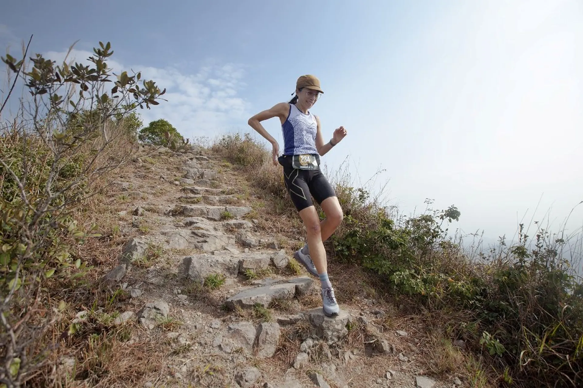 The image shows a person running or hiking down a rocky trail on a hillside. They are wearing athletic gear, including a hat, tank top, shorts, and a waist belt. The background features vegetation and a clear sky.
