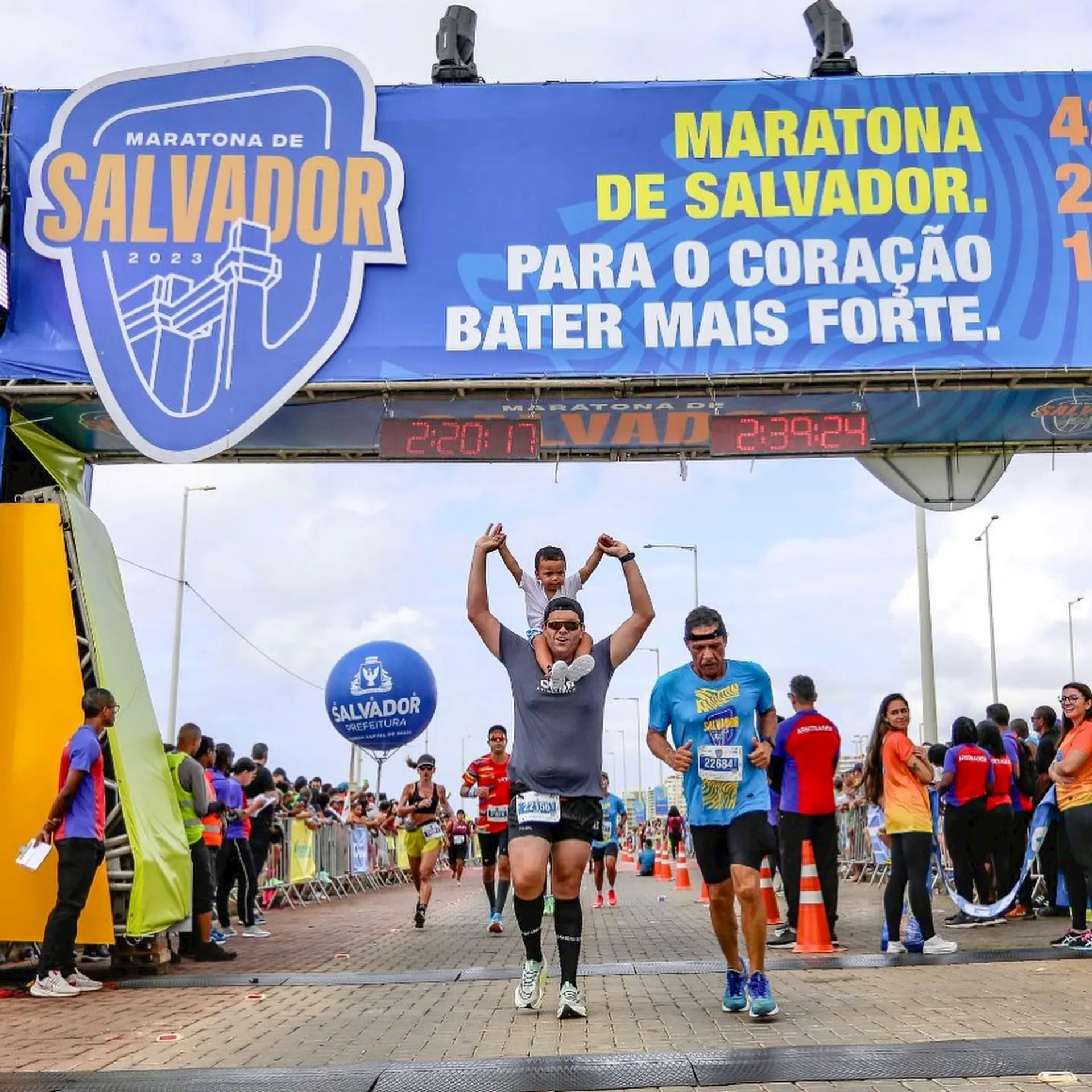 In the image, we see a runner crossing the finish line of a marathon. The event is called "Maratona de Salvador," as indicated by the banner above the finish line. The banner also includes the year, 2023, and a motivational slogan in Portuguese which translates to "For the heart to beat stronger." The runner is raising both arms in a gesture of triumph or celebration. Other participants can be seen in the background, along with spectators and race infrastructure like barriers and tents. The digital clock displays the time elapsed since the race began, suggesting this runner’s personal finish time or the time since the race officially started.