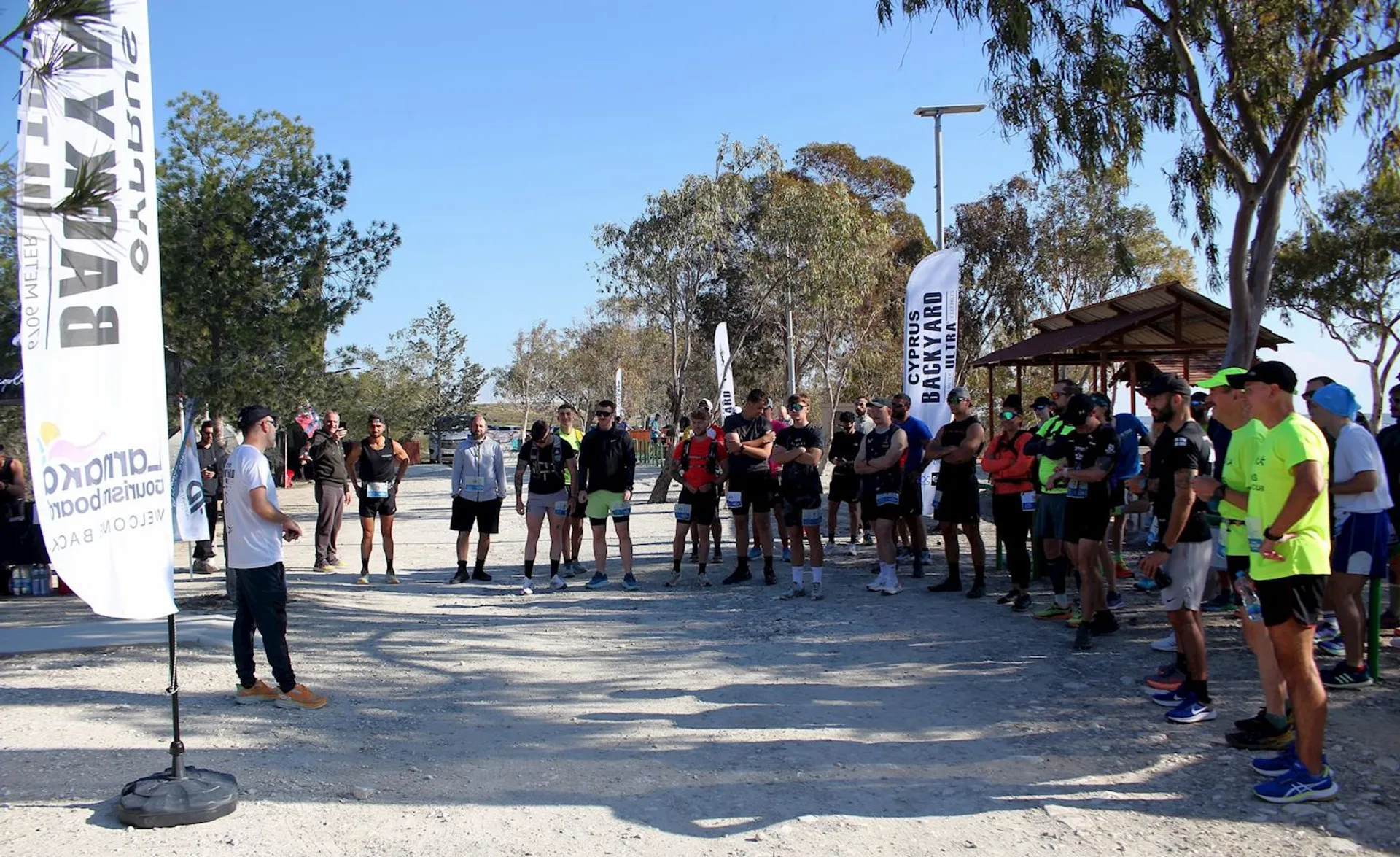 The image shows a group of people gathered outdoors, possibly for a running event or group activity. A person is standing in front of the group, possibly speaking or giving instructions. There are banners and trees visible in the background. The participants appear to be dressed in athletic clothing, suggesting a sporting or fitness-related event.
