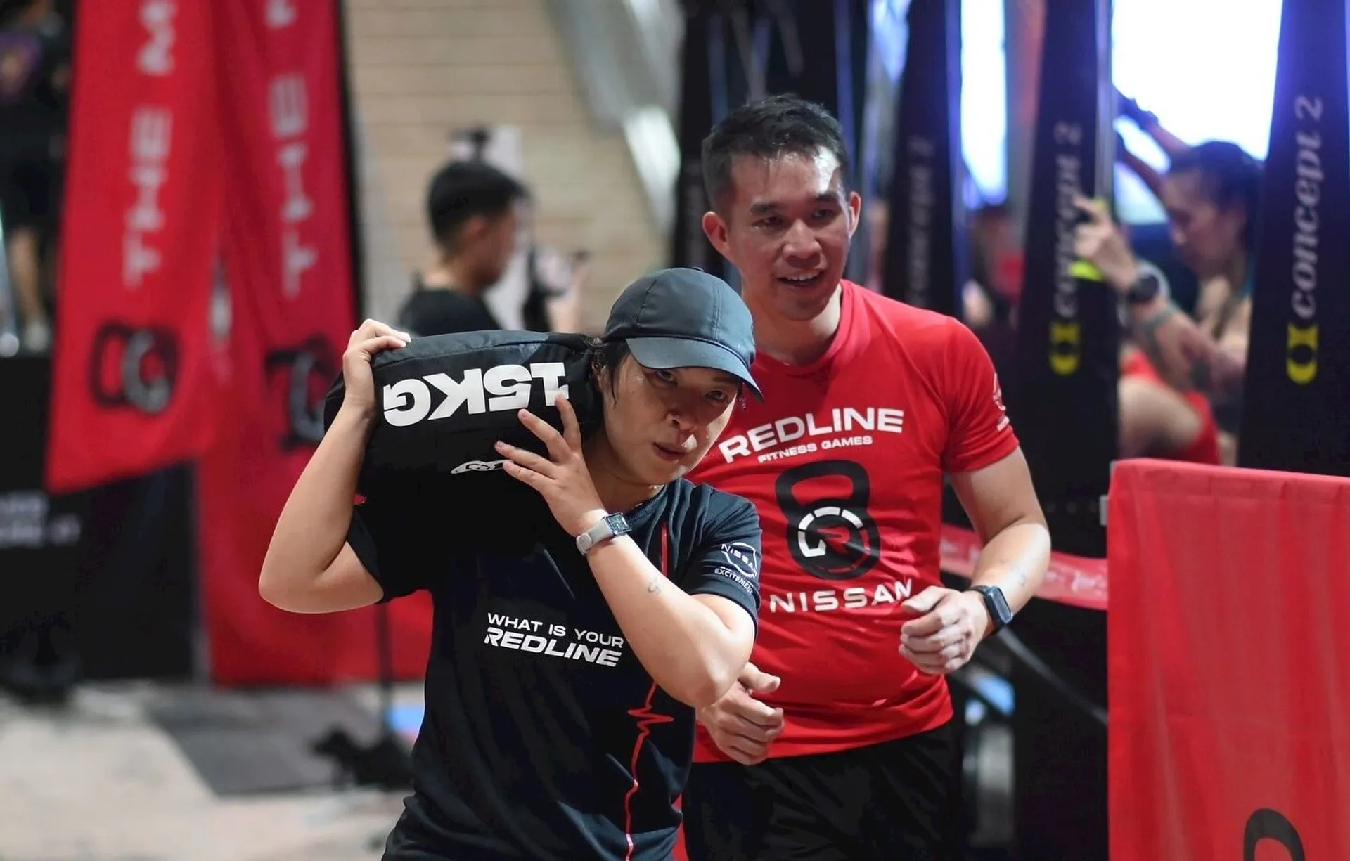 This image shows two people participating in a fitness event. The person in the foreground is carrying a sandbag, and both individuals are wearing athletic attire and have shirts with event branding. The setting seems to be an indoor gym or competition area.