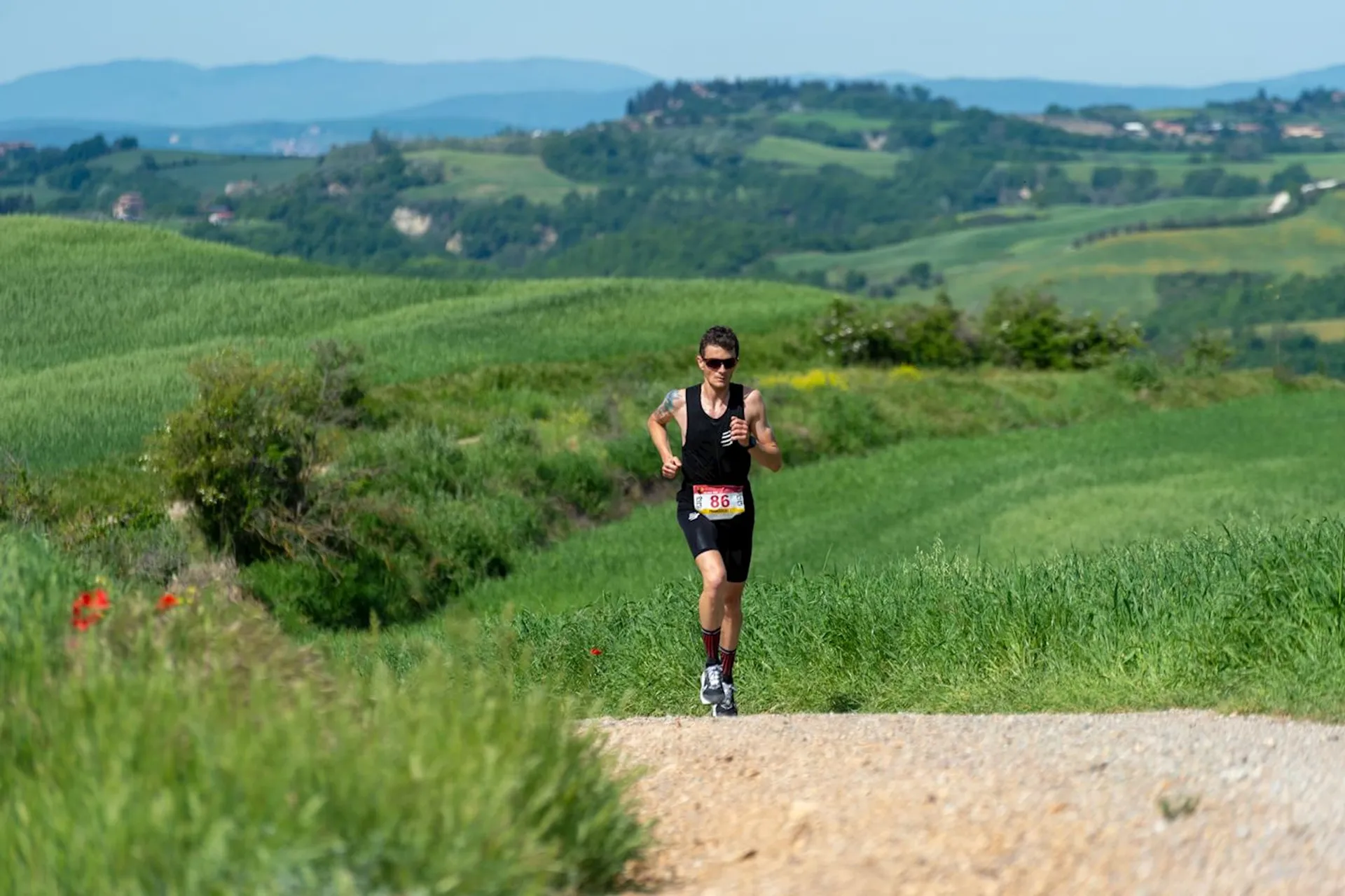 The image shows a person running on a gravel path through a lush, green landscape. They are wearing sports attire, sunglasses, and a running bib. The background consists of rolling hills and a clear blue sky.