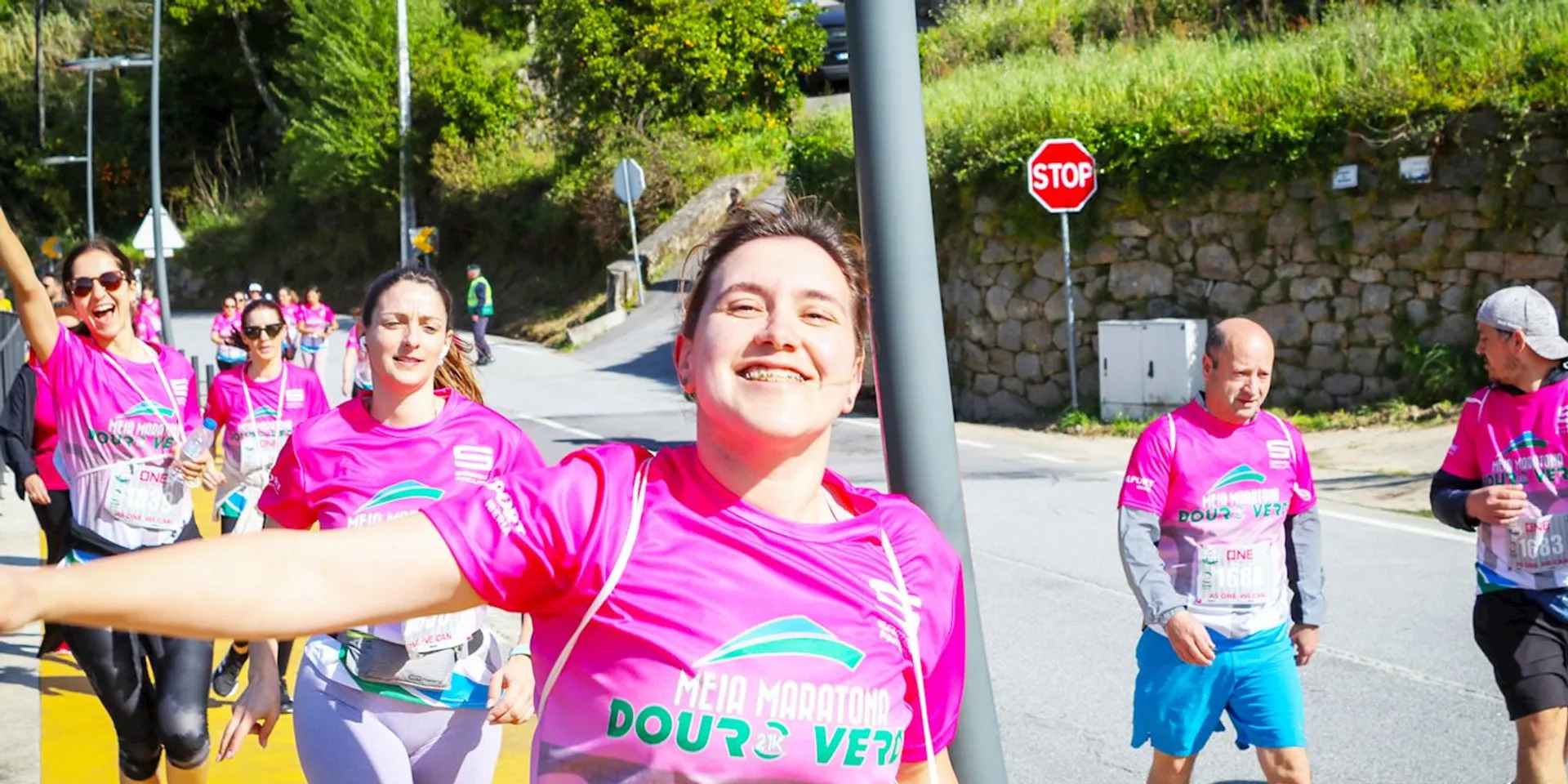 The image shows a group of participants in a running event, likely a half marathon as indicated by the text on their pink shirts. The central figure appears to be a happy and enthusiastic woman who is taking a selfie or video as she runs. Behind her are other equally energetic participants, with one woman wearing sunglasses and a headband. In the background, there is a stop sign and a sign that might indicate the name of the running event or the place where it is being held. It's a sunny day, and the setting seems to be a street with some greenery around. The overall atmosphere is one of enjoyment and physical activity.