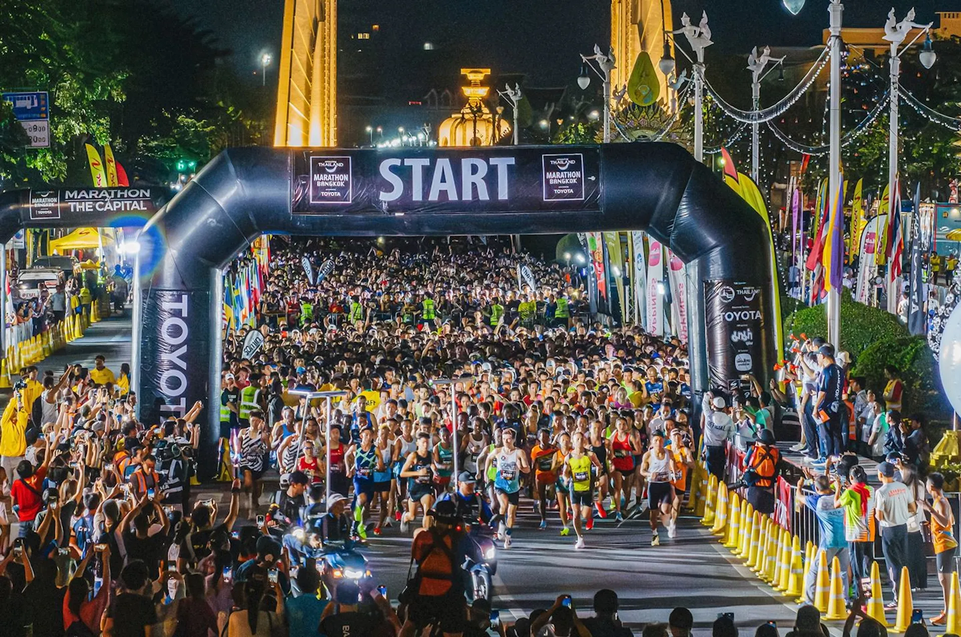 The image shows the start of a marathon or race event. There is a large crowd of runners at the starting line, and many spectators and photographers along the sides. The event appears to be taking place at night, as there are lights illuminating the area. There is a large inflatable arch with the word "START" on it, indicating the beginning of the race.