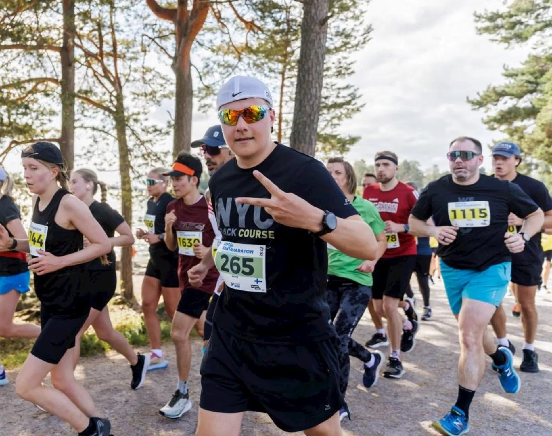 The image shows a group of people running in a race. They are outdoors and wearing numbered bibs on their chests. Some runners are wearing sunglasses and hats, and the setting appears to be a wooded area or park. The weather looks clear and sunny.