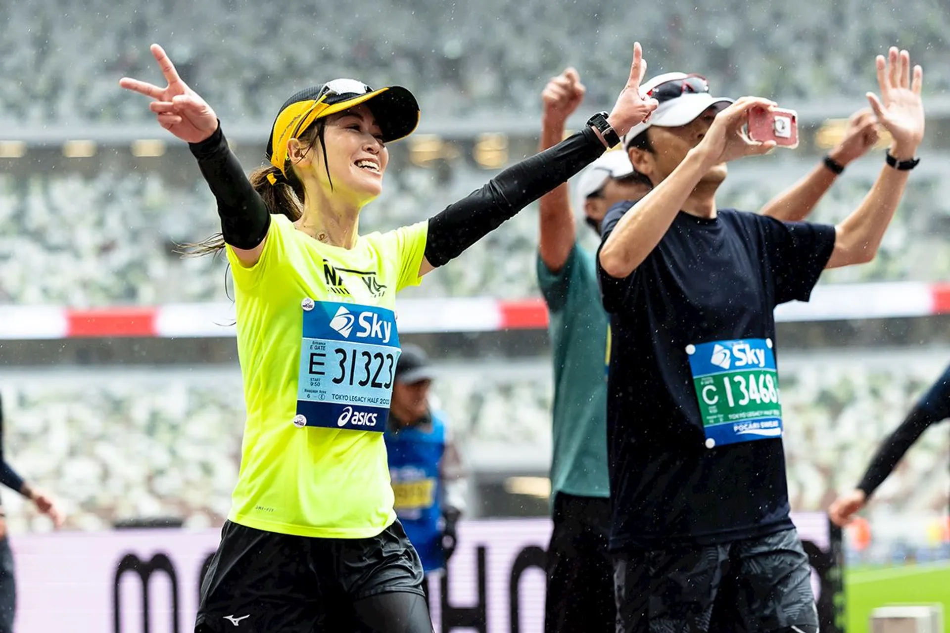 The image shows a marathon scene with runners celebrating. A woman in a yellow shirt is making a victory gesture with her arms raised. Another person next to her is taking a photo with a smartphone. Both are wearing race bibs. The background appears to be a stadium or race setting.