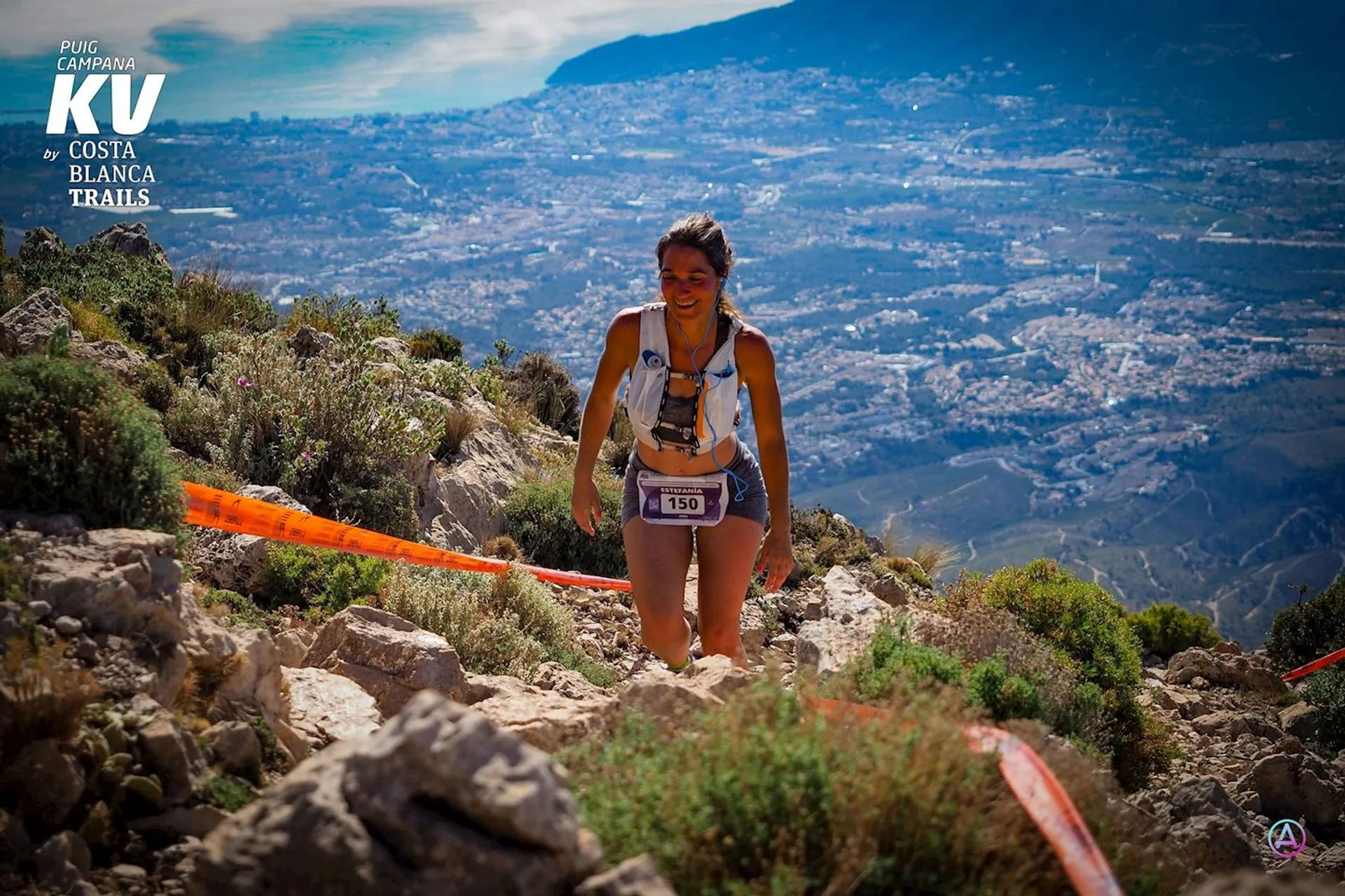 In the image, there's a woman running uphill on a rocky trail during a