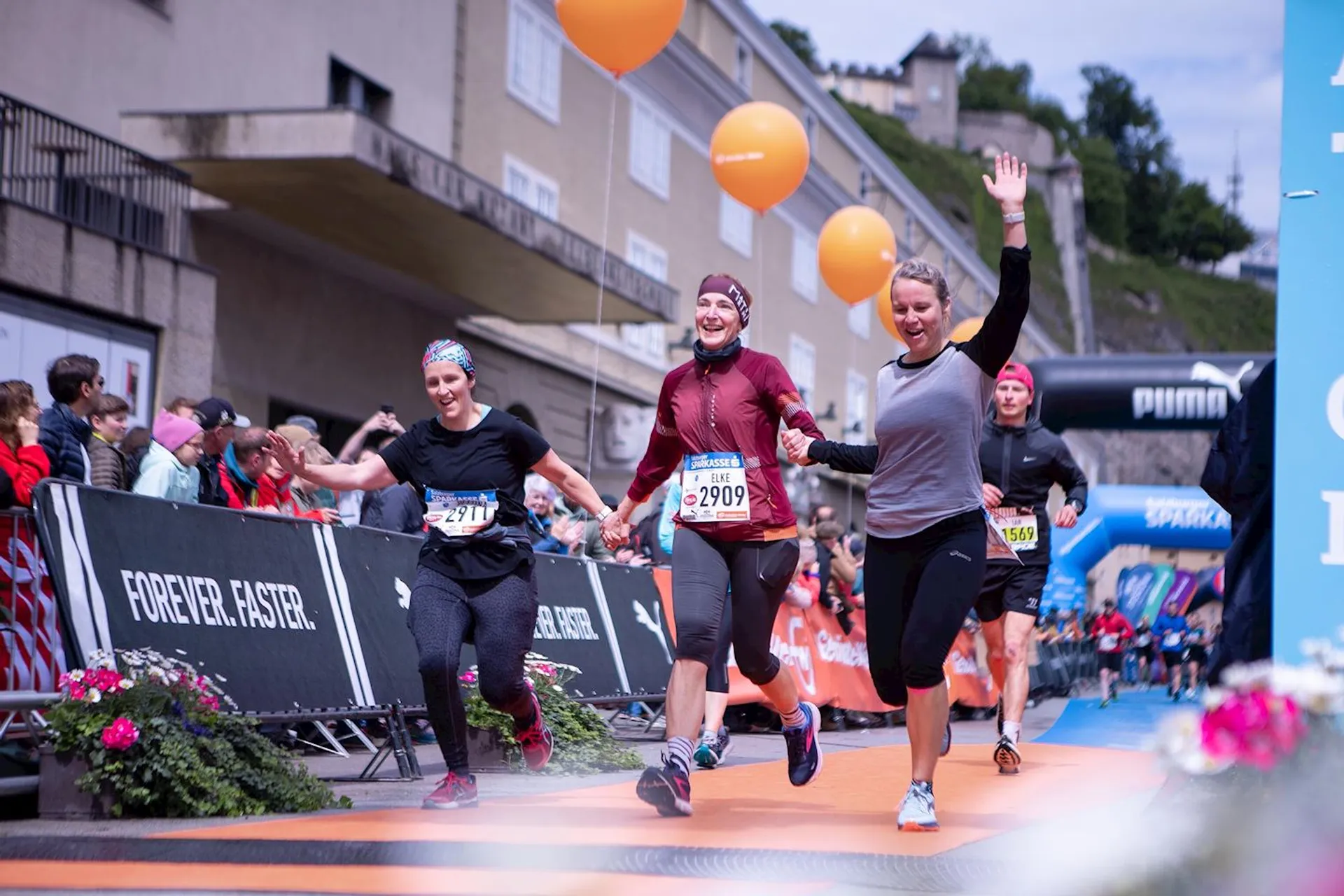 The image shows a group of runners crossing a finish line during a race. There are several people in the background, and the runners appear excited and happy. Some of them are holding up their arms in celebration. The scene has a few balloons and banners, and there are spectators on the sides.