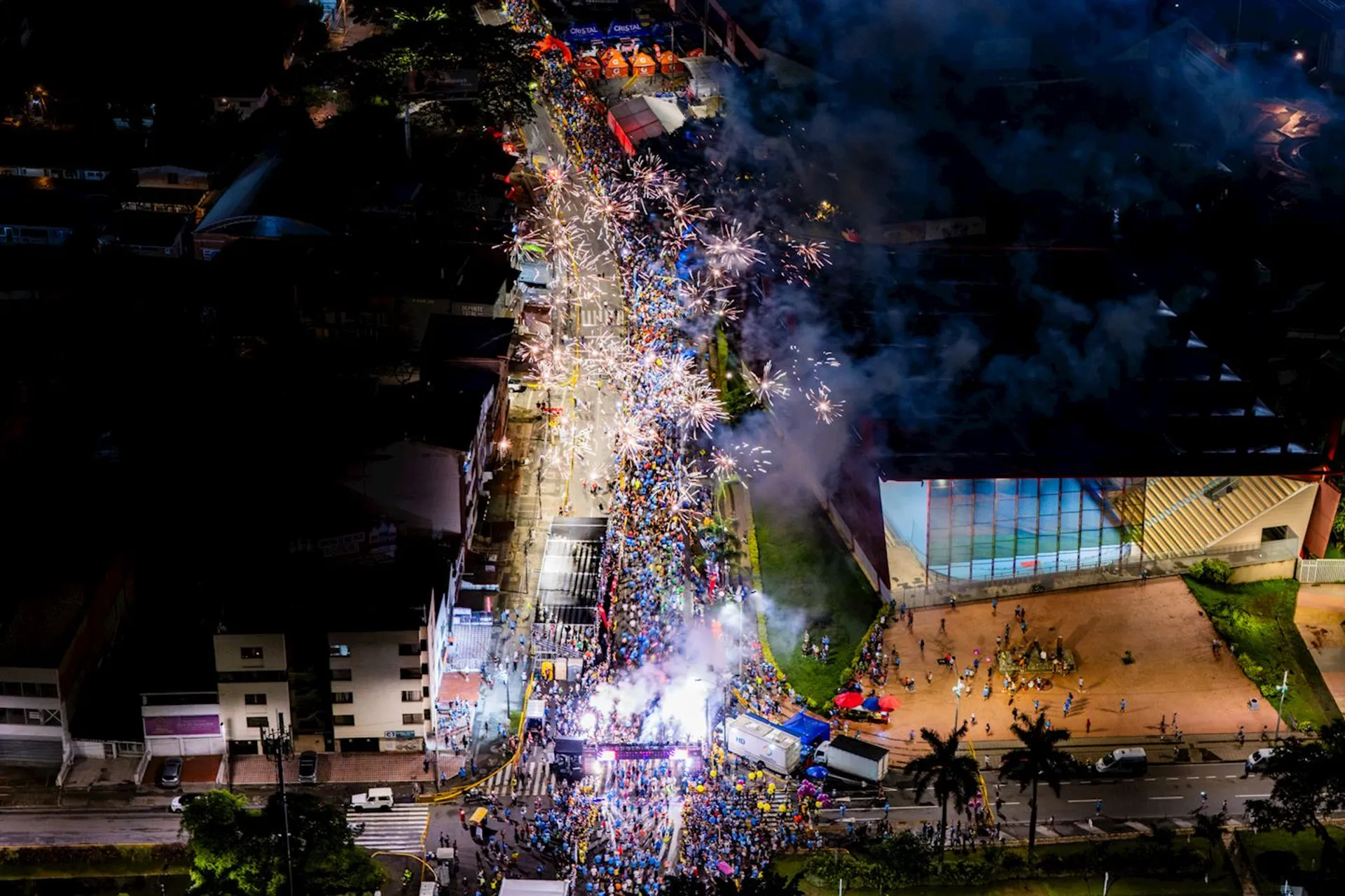 This image shows a vibrant street festival or parade at night. There's a large crowd of people gathered along a street, with bright lights, smoke, and fireworks creating a lively and festive atmosphere. Buildings and trees line the street, and a lit-up area appears to be a focal point of the celebration.