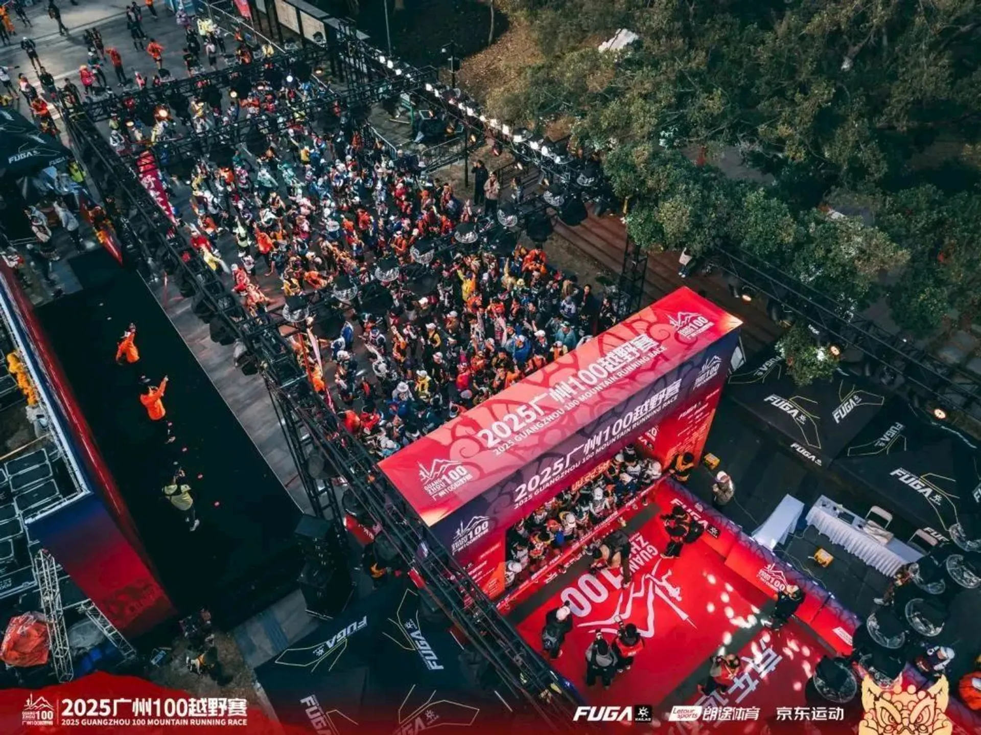 This image shows a large public event or gathering, likely a promotional or competitive event. It features a red stage with banners displaying the year "2025" and other details in Chinese. There is a crowd seated in front of the stage, with various lighting and equipment set up around the area. The setting appears to be an outdoor venue possibly related to a gaming or entertainment event, as indicated by the logos and decorations.