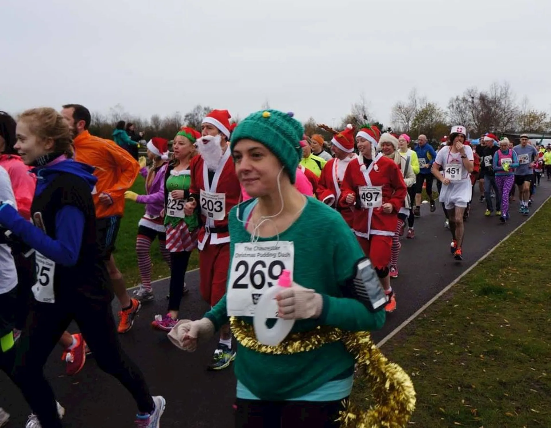 The image depicts a group of people participating in a fun run or similar event. Many of the participants are dressed in festive costumes, suggesting it may be a holiday-themed event, such as a Christmas or Santa run. Several individuals are dressed like Santa Claus, and others are wearing Christmas-themed outfits like elf costumes. The presence of race bibs on the participants indicates it's an organized running event. The setting appears to be a park or open space with trees in the background, and the weather seems overcast, as many of the participants are wearing long sleeves or jackets. Everyone seems to be in good spirits, enjoying the communal activity.