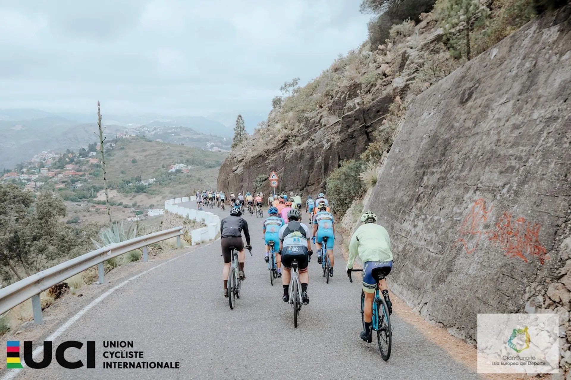The image depicts a cycling event, with a group of cyclists riding along a winding mountain road. The riders appear to be decked out in professional cycling gear, suggesting that this might be a part of a competitive race or an organized cycling tour. The terrain consists of a steep hillside, and it seems to be a cloudy day. The banners on the bottom left corner with the UCI (Union Cycliste Internationale) logo and other logos suggest this event is sanctioned by the international governing body for cycling.