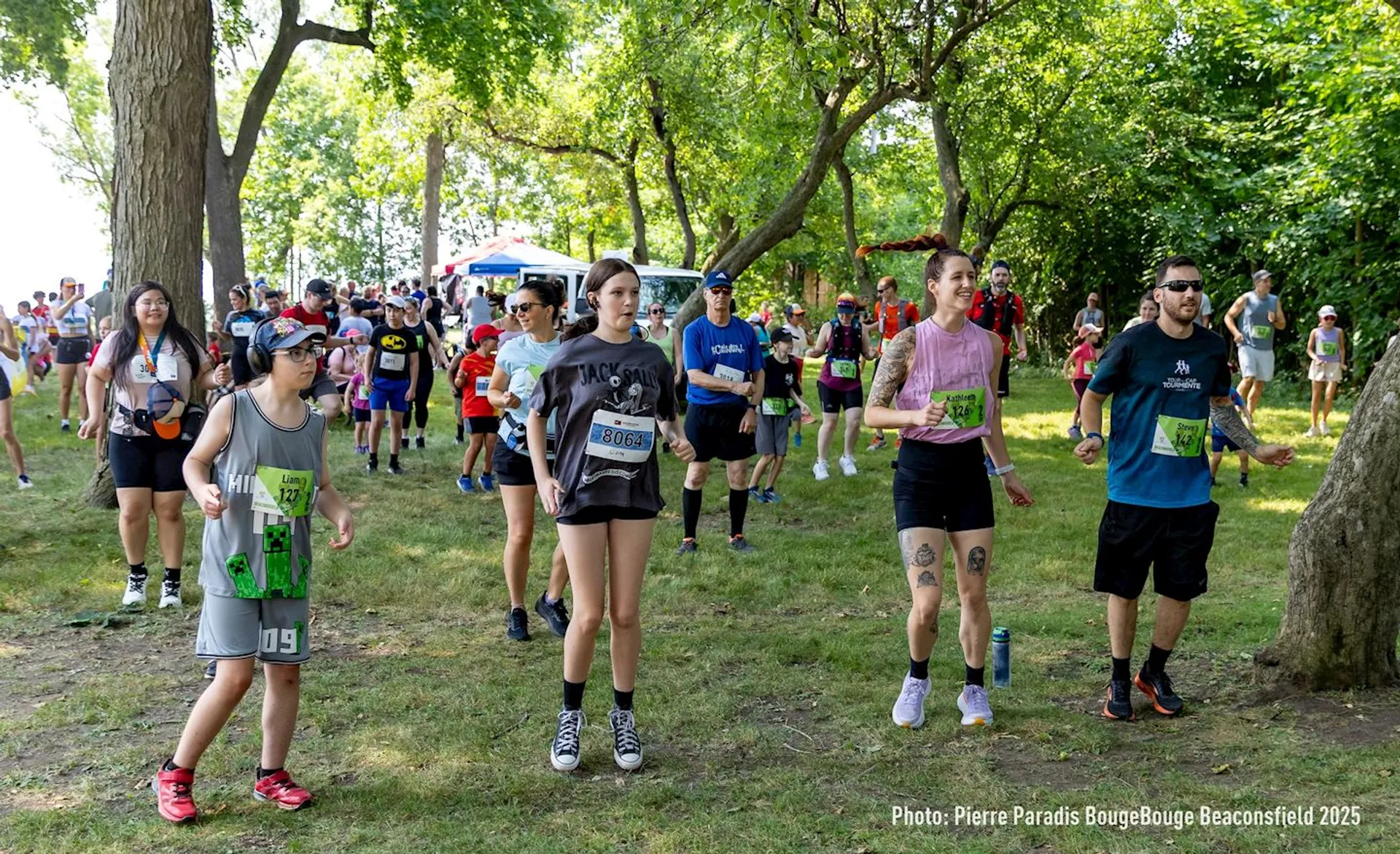 The image shows a group of people participating in an outdoor event, likely a fitness or running activity, given their athletic attire and the bib numbers some are wearing. They are in a grassy, tree-lined area, which suggests a park or natural setting. Some participants are stretching or warming up, and there's a mix of adults and children present.