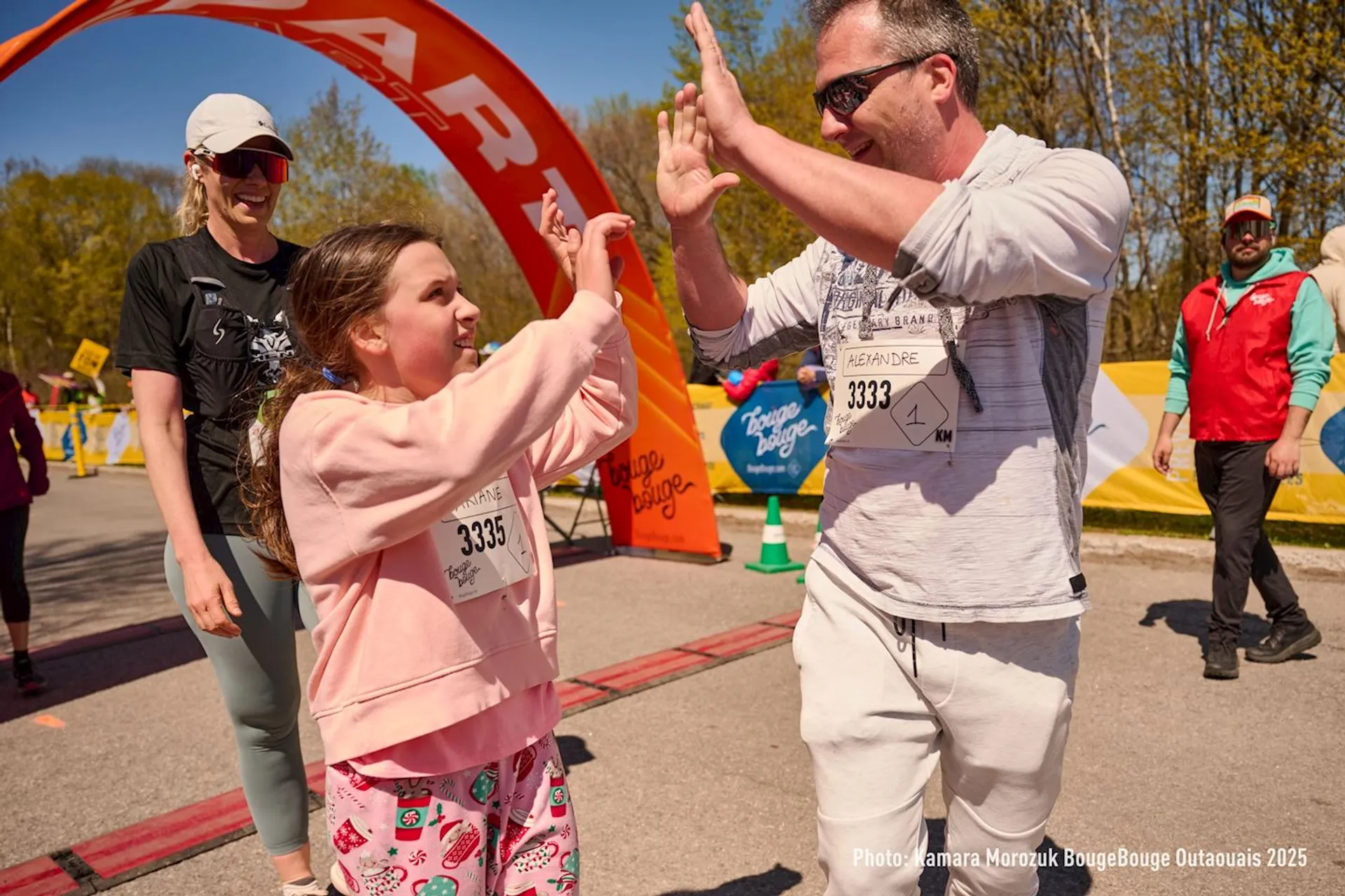 The image shows a group of people at what appears to be a running event or race. A man and a young girl are giving each other a high five. They are wearing race bibs. Other participants and a staff member are visible in the background, along with a finish line banner and some trees.