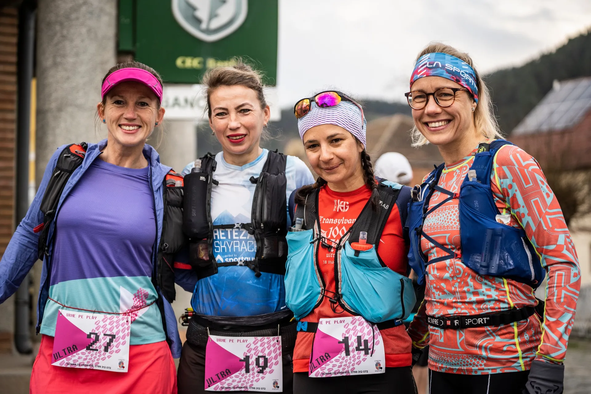 The image shows four runners or participants in a sporting event, likely a race, based on their athletic gear and bib numbers. Each person is wearing a running outfit with hydration packs. The bib numbers suggest they are part of an organized event. They are outdoors, and all seem to be smiling or posing for the photo.