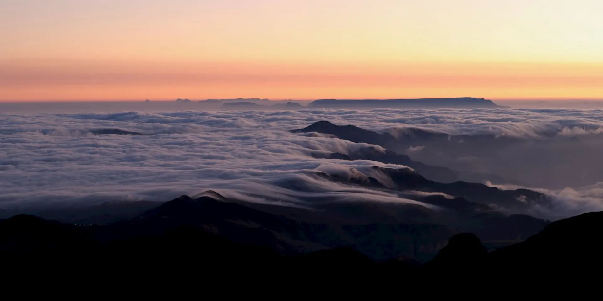 This image depicts a stunning landscape view at sunrise or sunset. It shows mountains with a layer of clouds cascading over them. The sky transitions from a warm orange to a soft pink and purple, casting a serene and ethereal atmosphere over the scene.