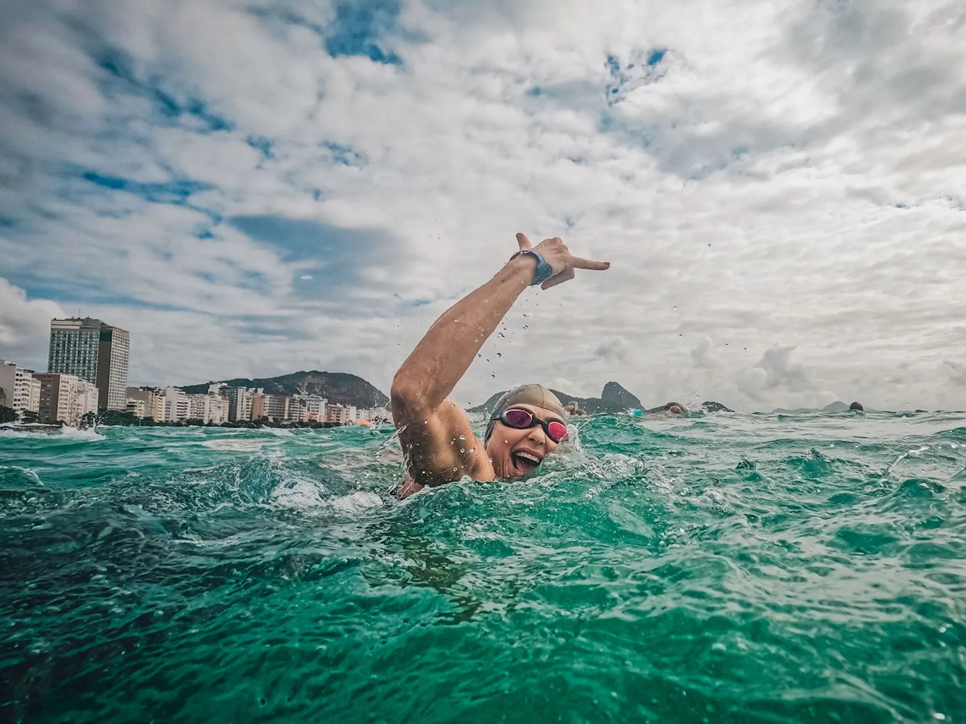 The image shows a swimmer in the ocean, wearing goggles and a swim cap, making a gesture with one hand. The background features a city skyline with tall buildings and distant mountains under a cloudy sky. It appears to be a vibrant and active scene.