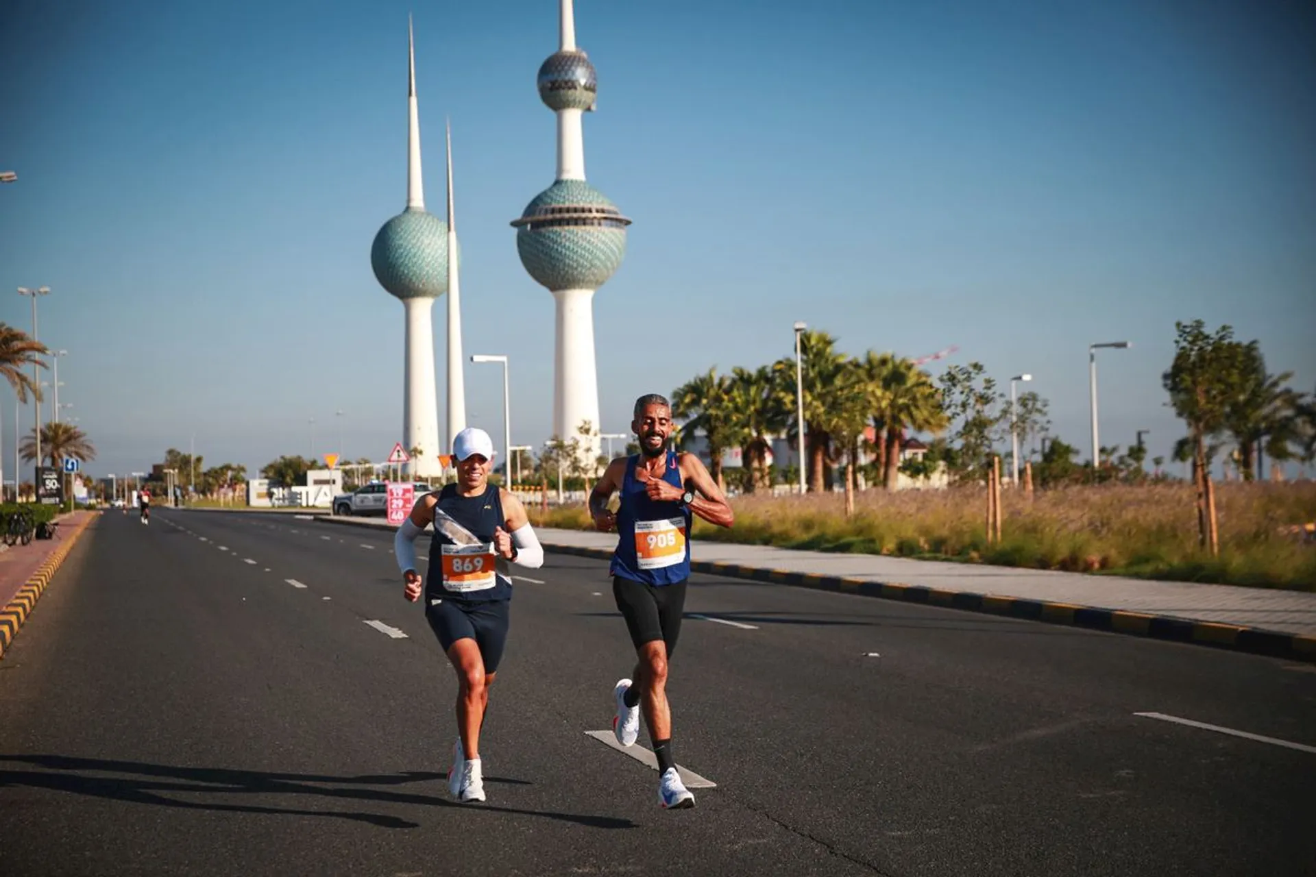 The image shows two runners on a road, with bib numbers 849 and 908, participating in what seems to be a marathon or race. In the background, there are iconic towers, which appear to be the Kuwait Towers. The road is flanked by palm trees, and the weather looks clear and sunny.