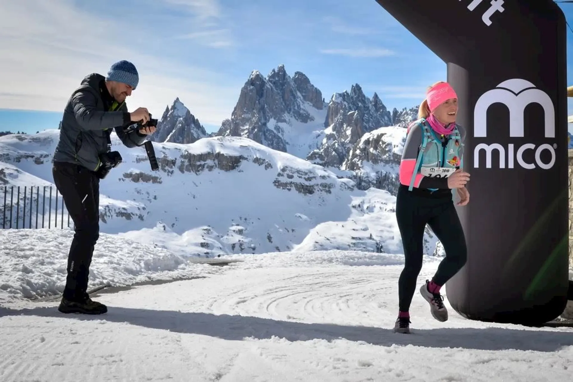 The image depicts a snowy mountain scene with two individuals prominently featured. On the left, there's a person holding a camera, taking a photo or filming. This person is wearing winter clothing, including a beanie and gloves, suitable for the cold environment. On the right, there is another individual who seems to be participating in a winter sports event, wearing a number tag and athletic attire, and is likely running towards a finish line indicated by a branded archway. The person is also wearing a beanie and sunglasses, indicating that they are actively engaged in the sport despite the cold temperatures. In the background, the jagged peaks of the mountains rise sharply against the clear blue sky, and the ground is covered with snow, emphasizing the alpine setting