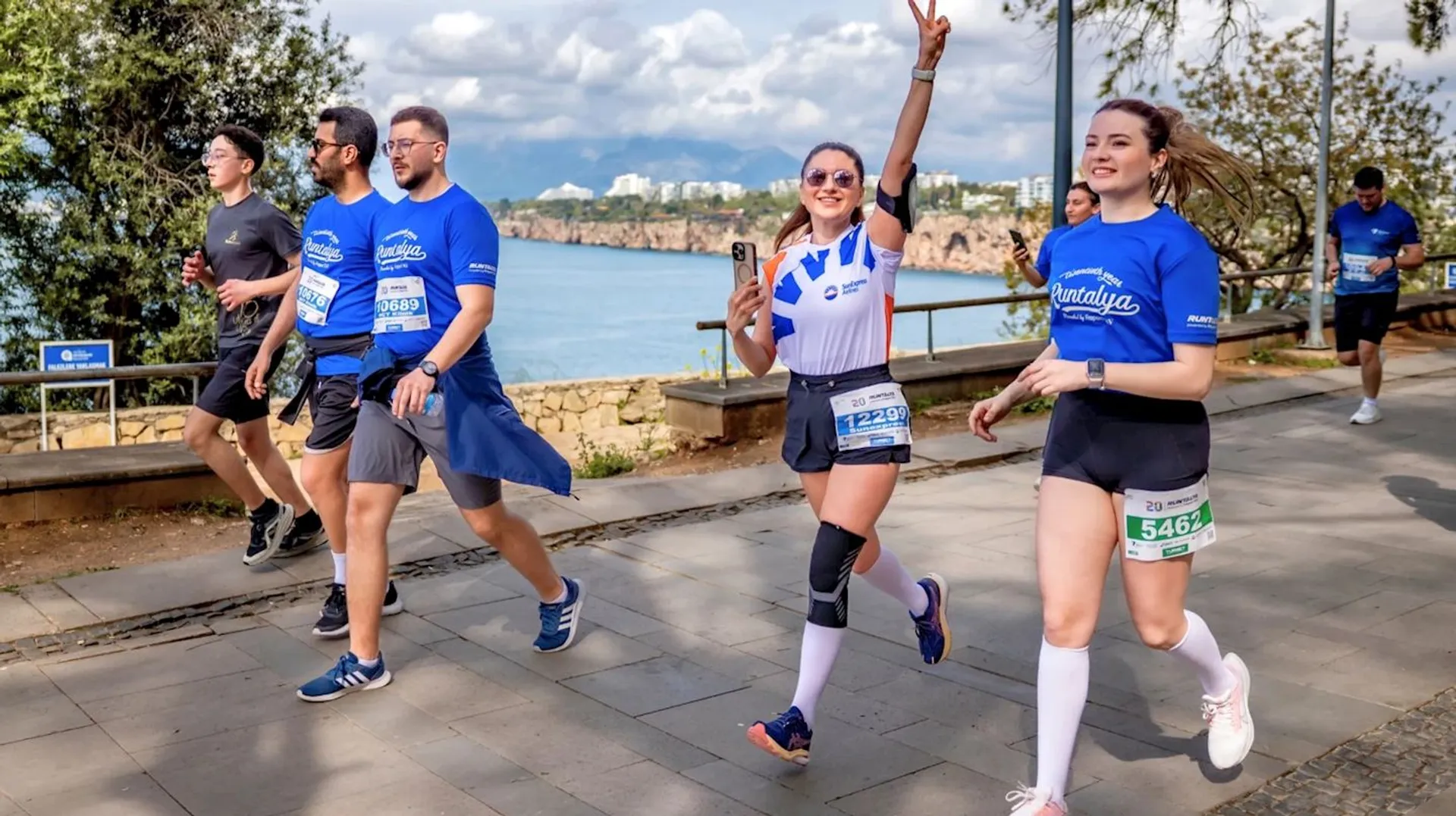 The image shows a group of people participating in a walking or running event. They are outdoors, walking on a paved path next to some water. The participants are wearing athletic clothing and bib numbers, suggesting it's a race or charity event. One person is posing with a peace sign and smiling at the camera. The surroundings include trees and a scenic view.
