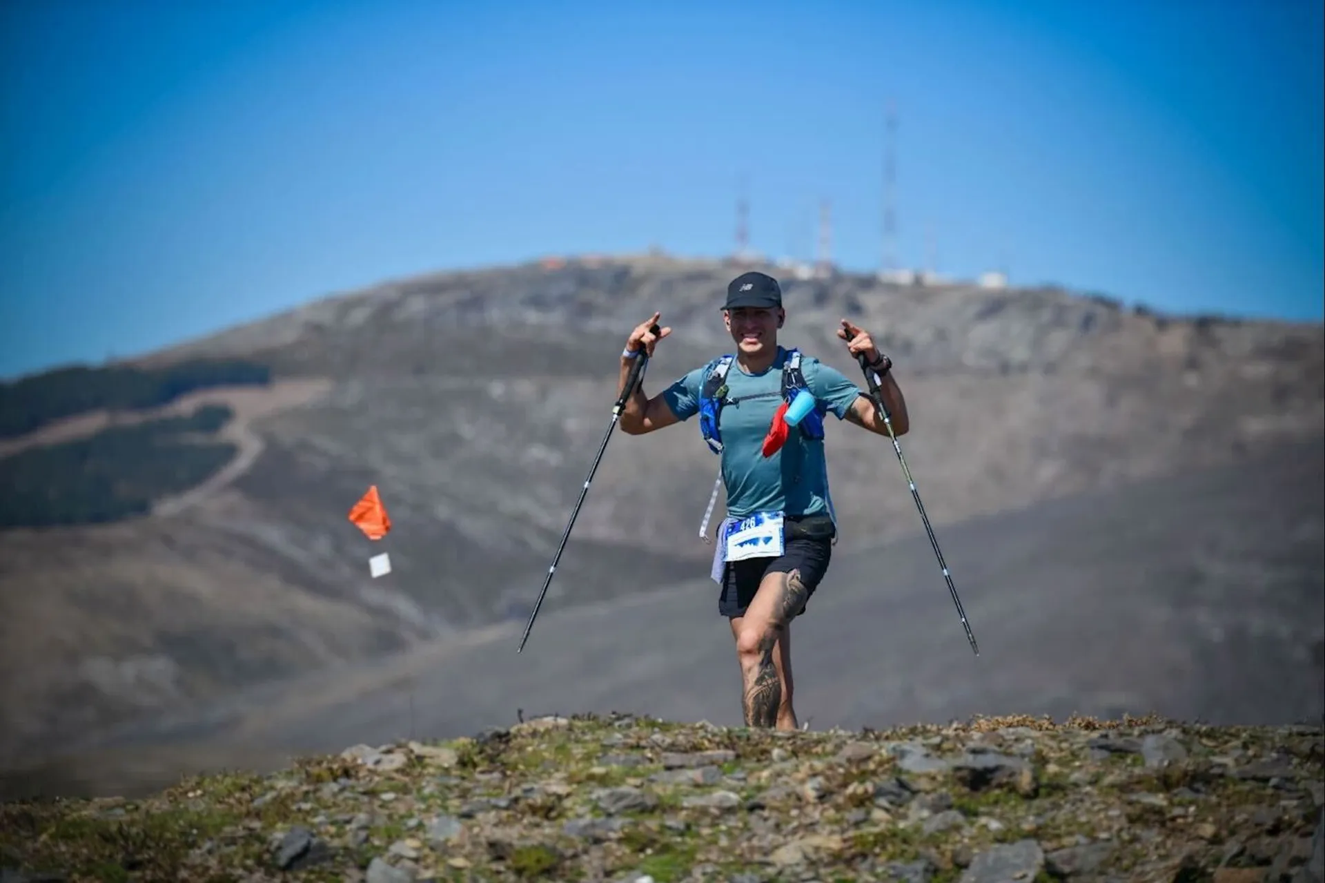 The image shows a person running on a rocky trail in a mountainous area. They are holding trekking poles and wearing a blue shirt, shorts, and a cap. There are small flags in the background, likely marking the trail, and the sky is clear and blue.