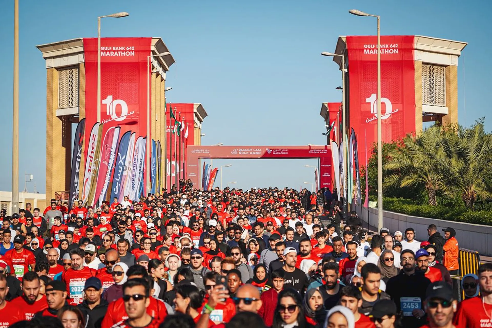 The image shows a large group of people participating in a marathon event. They are running on a wide road lined with flags and banners. Most participants are wearing red shirts, and the atmosphere conveys a sense of excitement and activity. The location appears organized, with structures set up for the event.