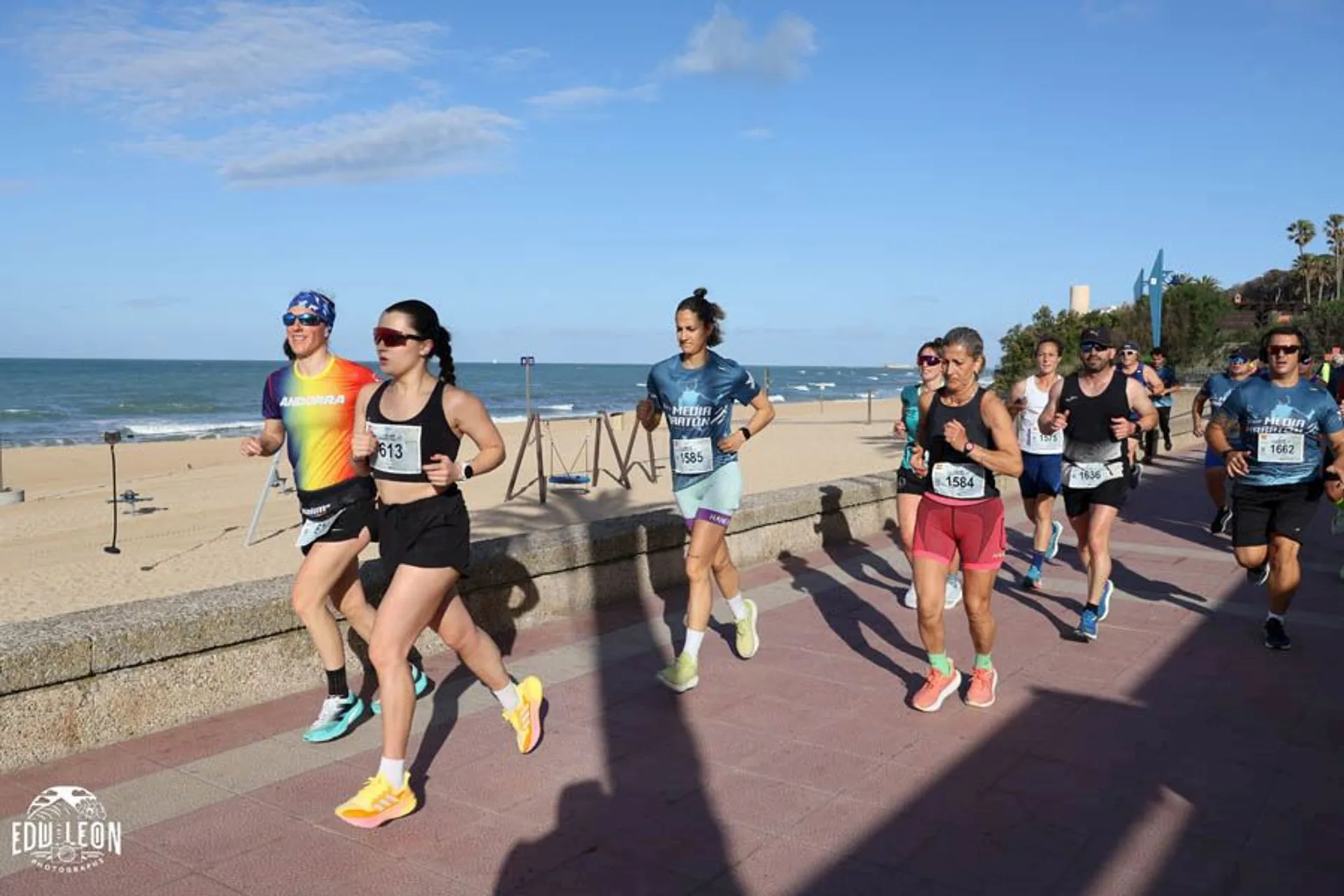 The image shows a group of people running along a paved path next to a beach. They are participating in a race or organized run, wearing athletic clothing and numbered bibs. The ocean is visible in the background under a clear blue sky.