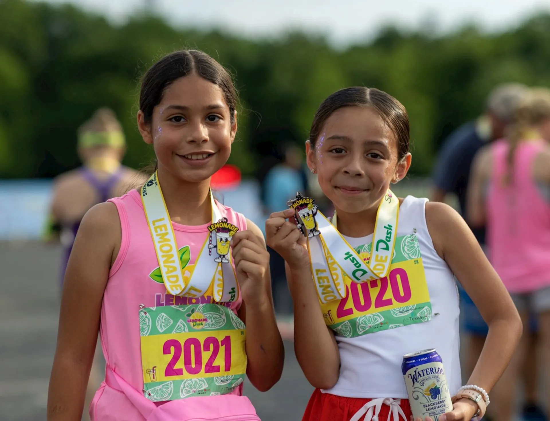 The image shows two children who have participated in a race. They are wearing bibs with the numbers 2021 and 2020, and holding medals. The bibs are labeled "LEMONADE" and they appear to be smiling. One of them is holding a can as well.