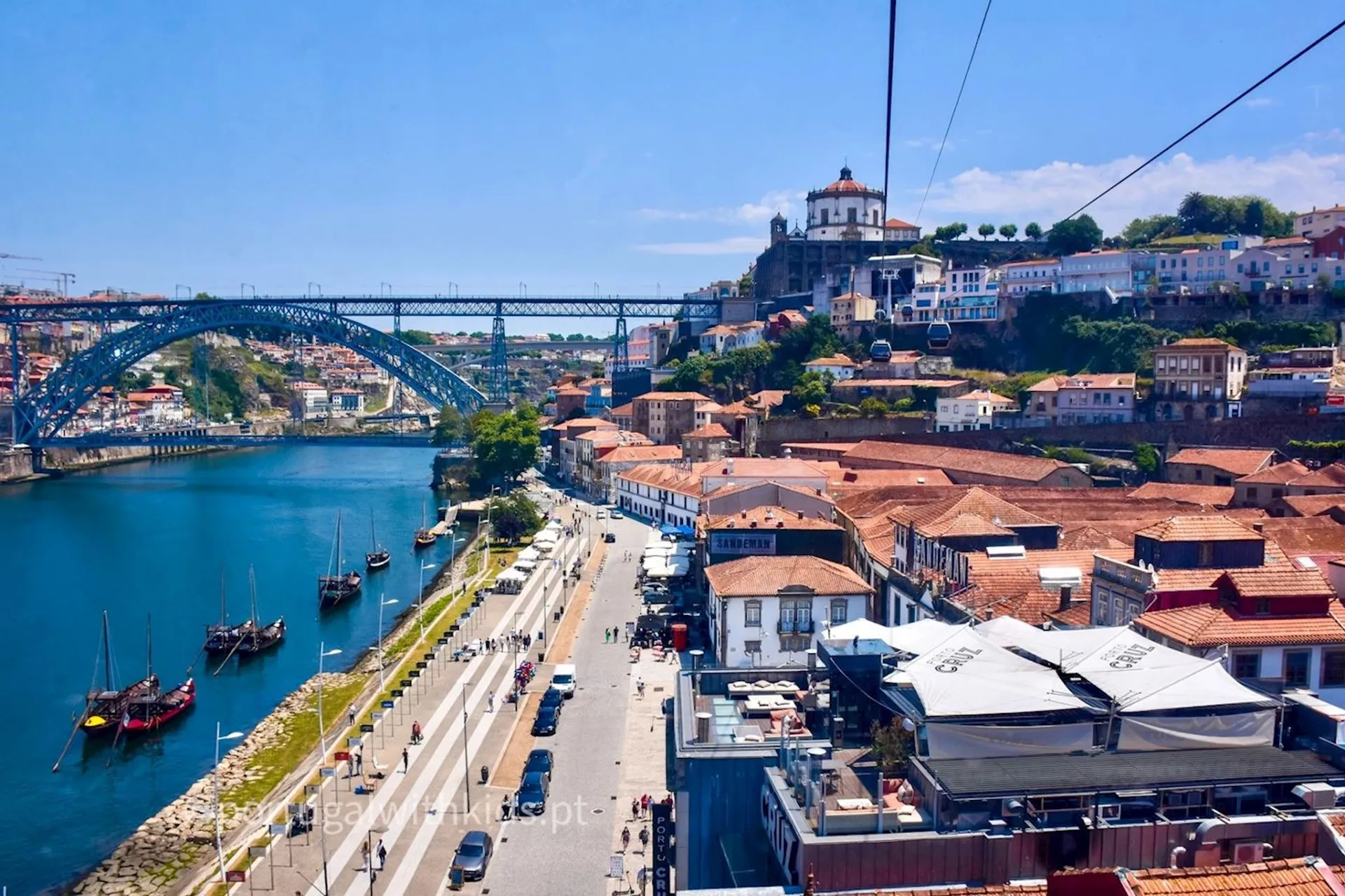 The image shows a beautiful panoramic view of a cityscape taken from an elevated perspective, possibly from a cable car or a high vantage point. It features a picturesque river scene with boats, a historic arch bridge to the left, and colorful buildings lining the waterfront. The architecture and the bridge suggest this could be a European city. There is a church or cathedral visible on the hilltop in the background, and the clear blue sky suggests a sunny day, perfect for enjoying the riverside promenade visible in the lower part of the image. The terracotta roof tiles and classical architectural style give the city a charming, timeless atmosphere.