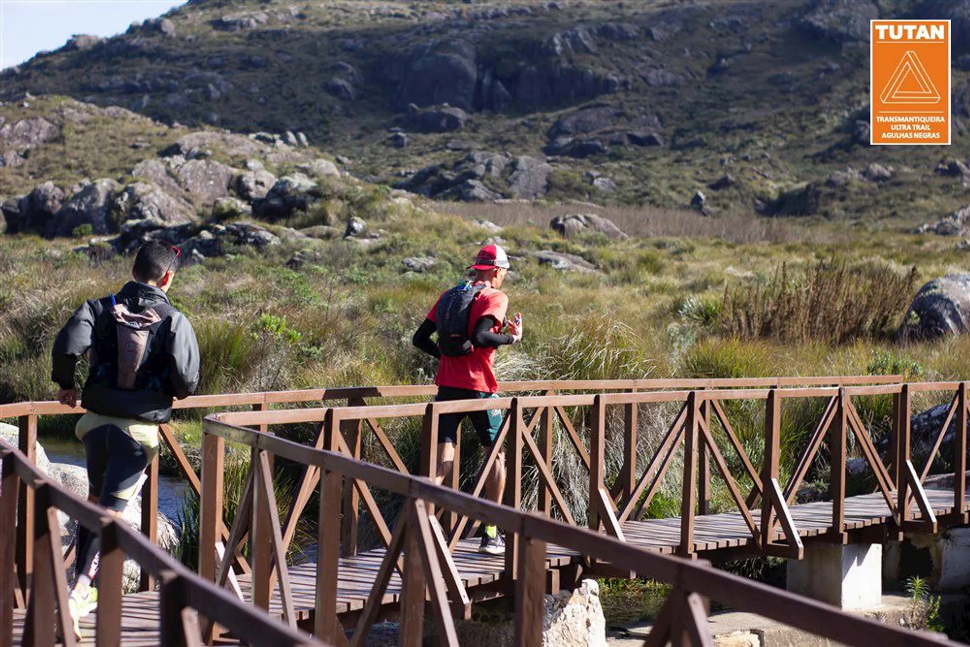 The image shows two individuals walking across a wooden bridge in a natural, rocky environment