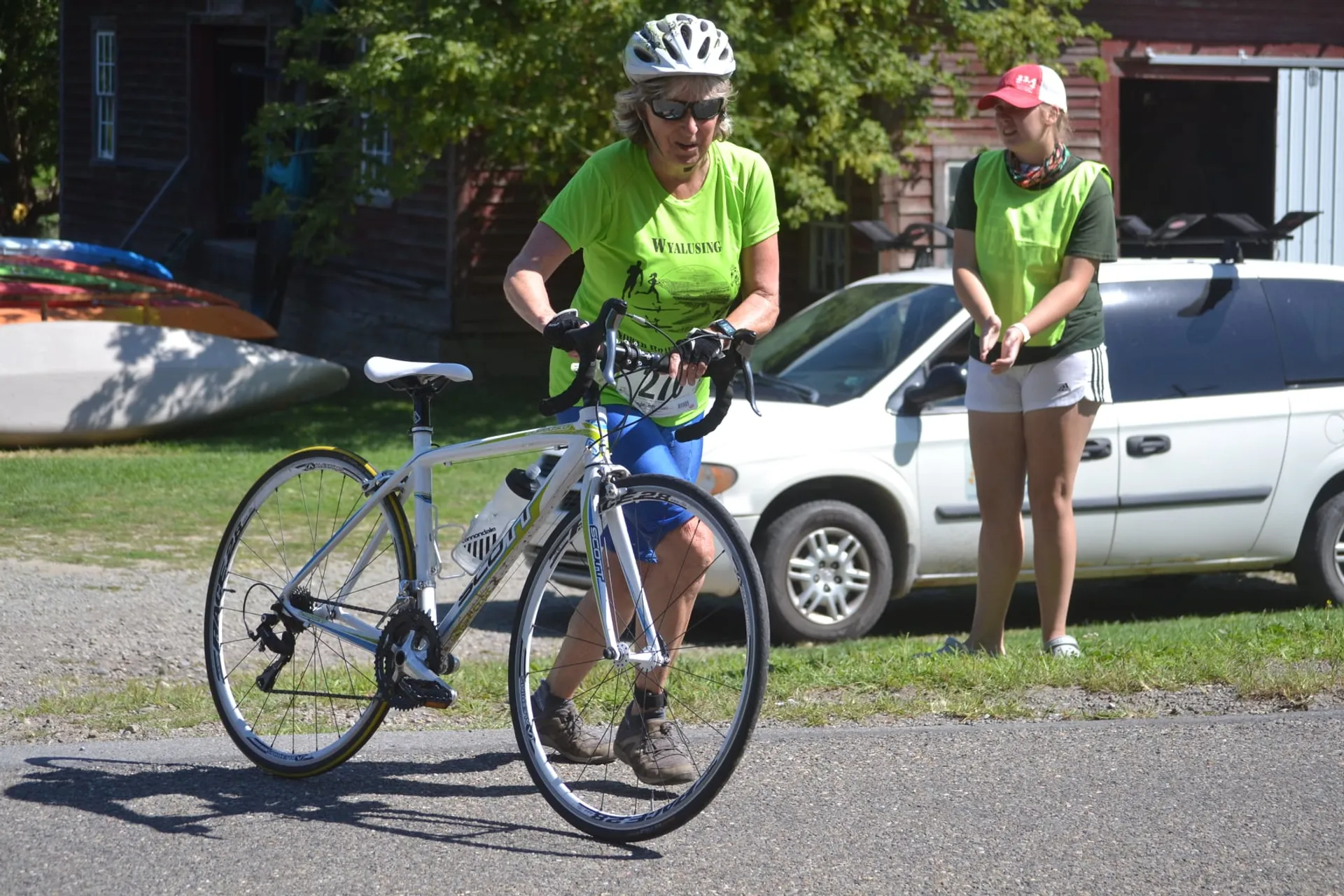 The image shows a person with a bicycle. The person is wearing a bright yellow shirt with some text on it, a helmet, sunglasses, gloves, and cycling shorts. They seem to be preparing for a bike ride or taking a short break as the bike is stationary and they are standing beside it while holding onto the handlebars.

Behind the cyclist, there is a second person wearing a pink cap and a white shirt with a logo, shorts, and casual shoes. They appear to be looking at something off to the side and standing on what looks like a grassy area.

In the background, there's a minivan parked, and a canoe mounted on a rack on top of the vehicle. There are trees and a clear sky, suggesting that the