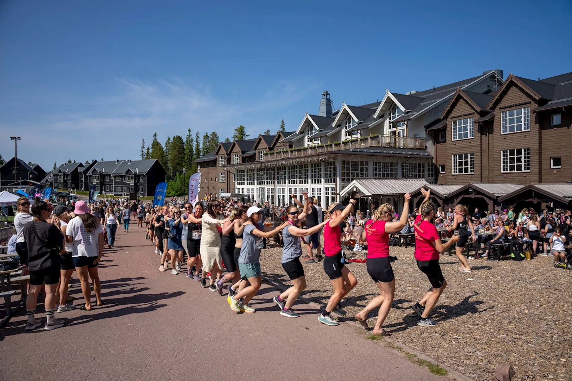 The image shows a group of people engaged in a line dance outdoors in front of buildings that resemble a resort or lodge. The participants appear to be having fun, with several onlookers and other groups around them. The setting is during the day, under clear blue skies.