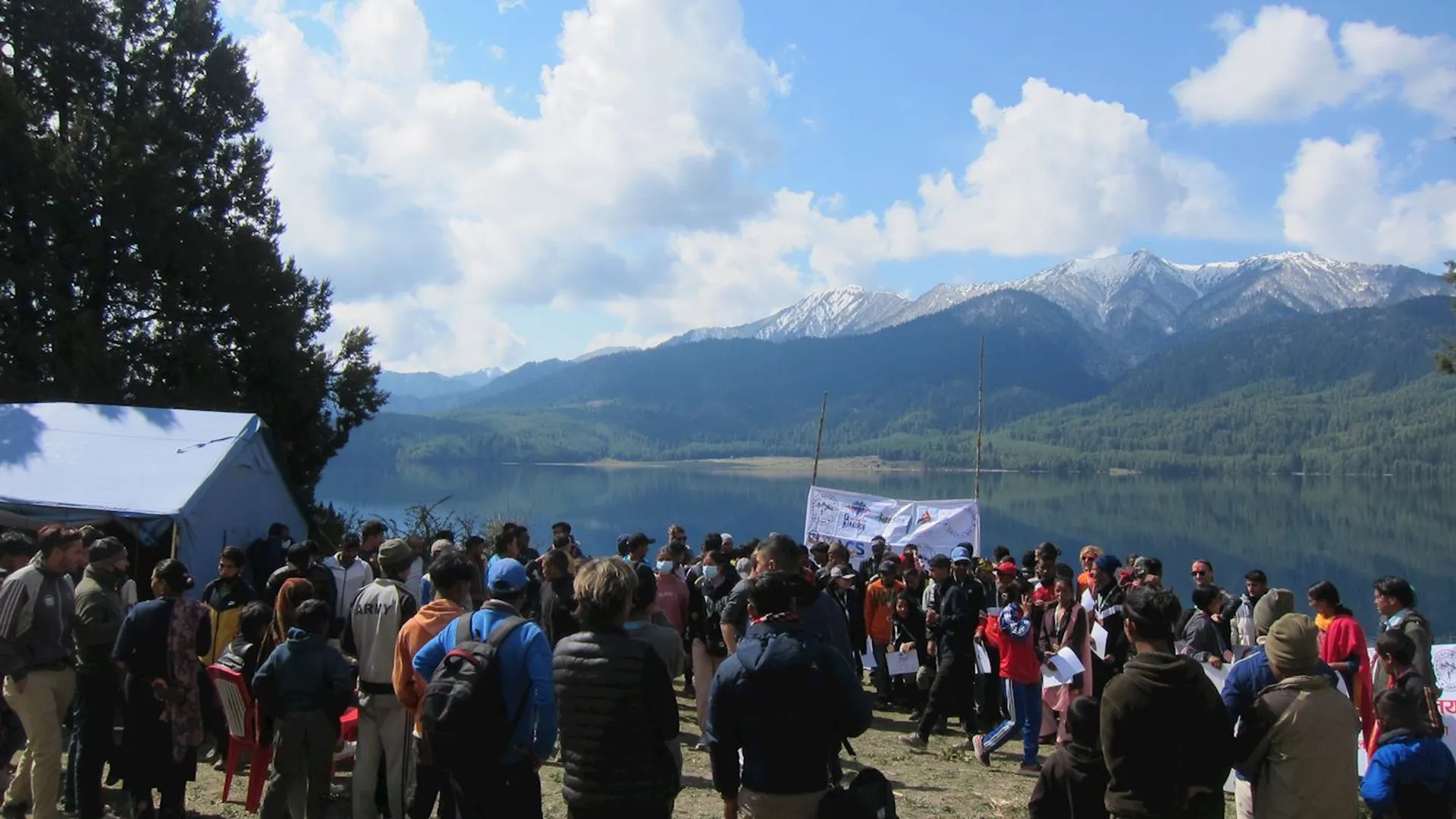 The image shows a group of people gathered outdoors near a lake with mountainous scenery in the background. There is a tent and a banner present, suggesting an organized event or gathering. The sky is partly cloudy, and the scene appears to be in a natural, scenic setting.