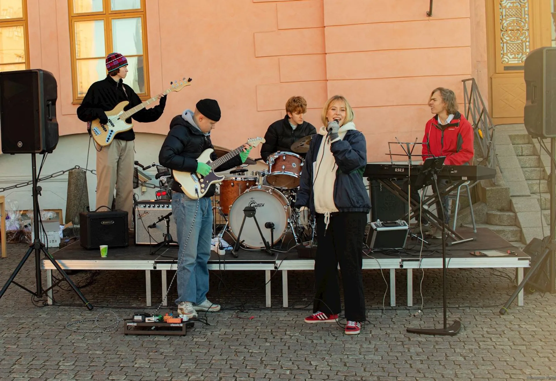 The image shows a group of musicians performing outdoors. There are five individuals visible,
