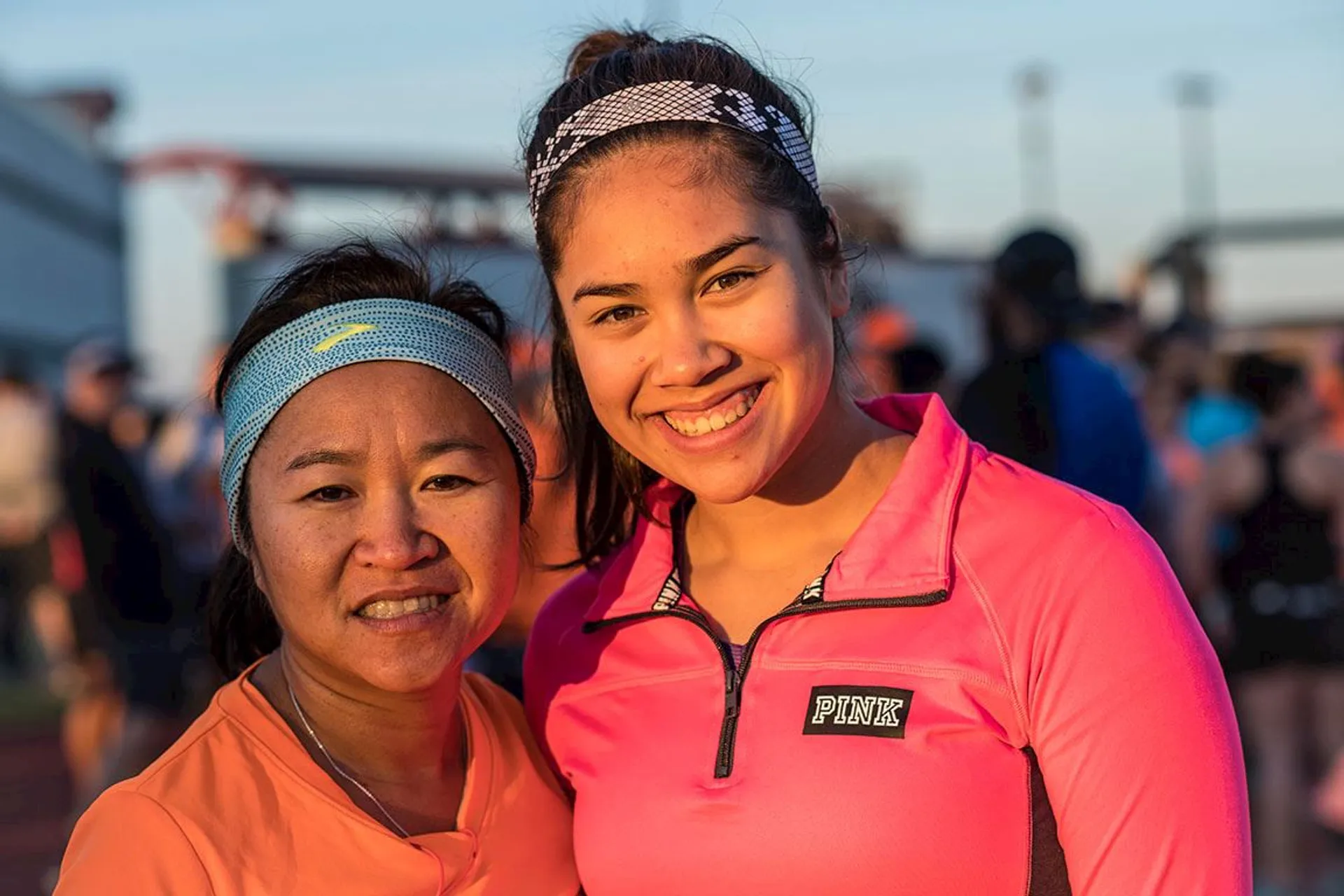 The image shows two people smiling at the camera. One is wearing an orange top and a headband, while the other is wearing a bright pink top with "PINK" written on it and a headband. They appear to be outdoors, possibly at an event or gathering, as there are other people in the background.