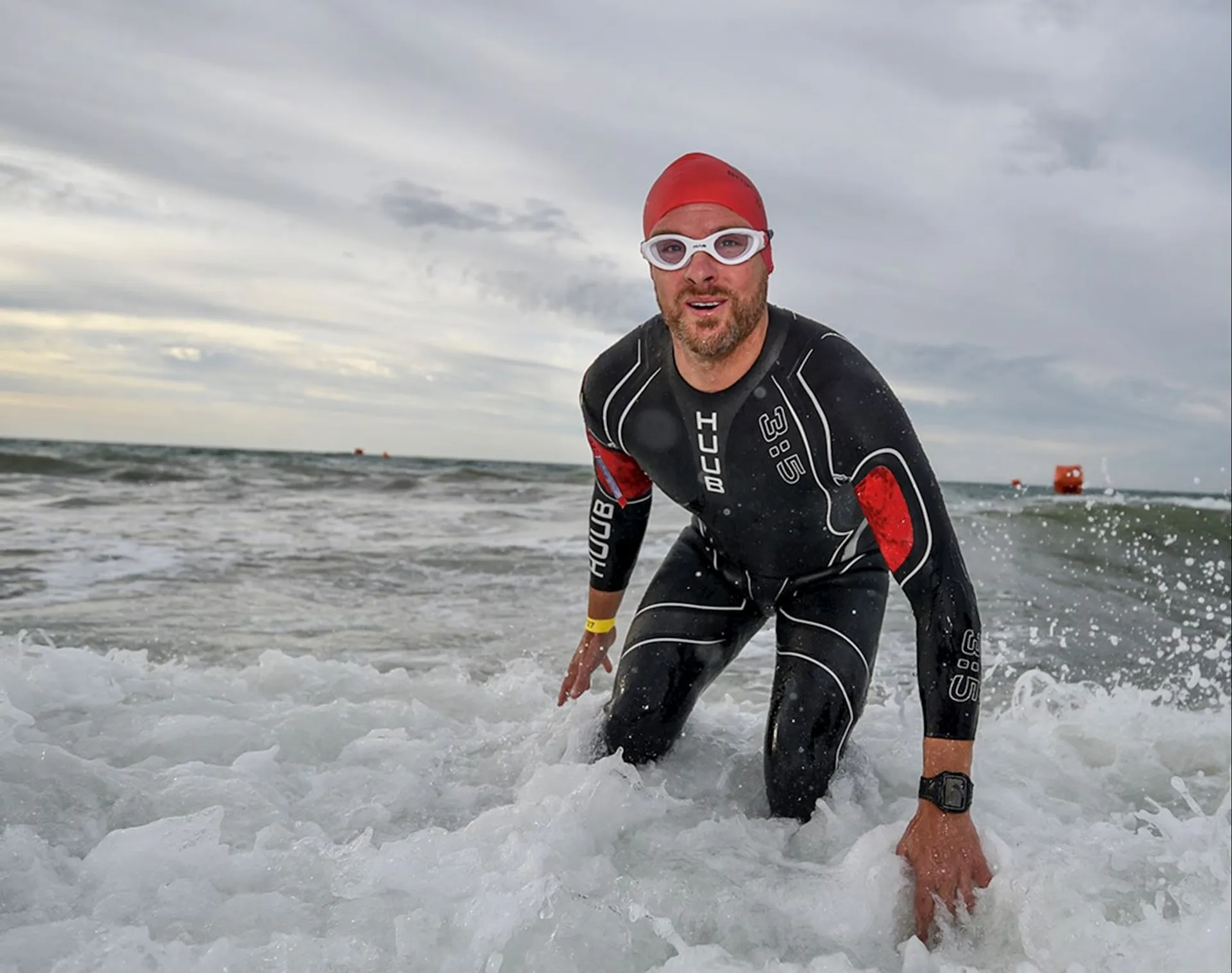 The image shows a person in a wetsuit, wearing a swim cap and goggles, coming out of the ocean water. It's likely part of a triathlon or a similar athletic event. The person is kneeling in the shallow water, and the background includes a cloudy sky and a buoy in the distance.