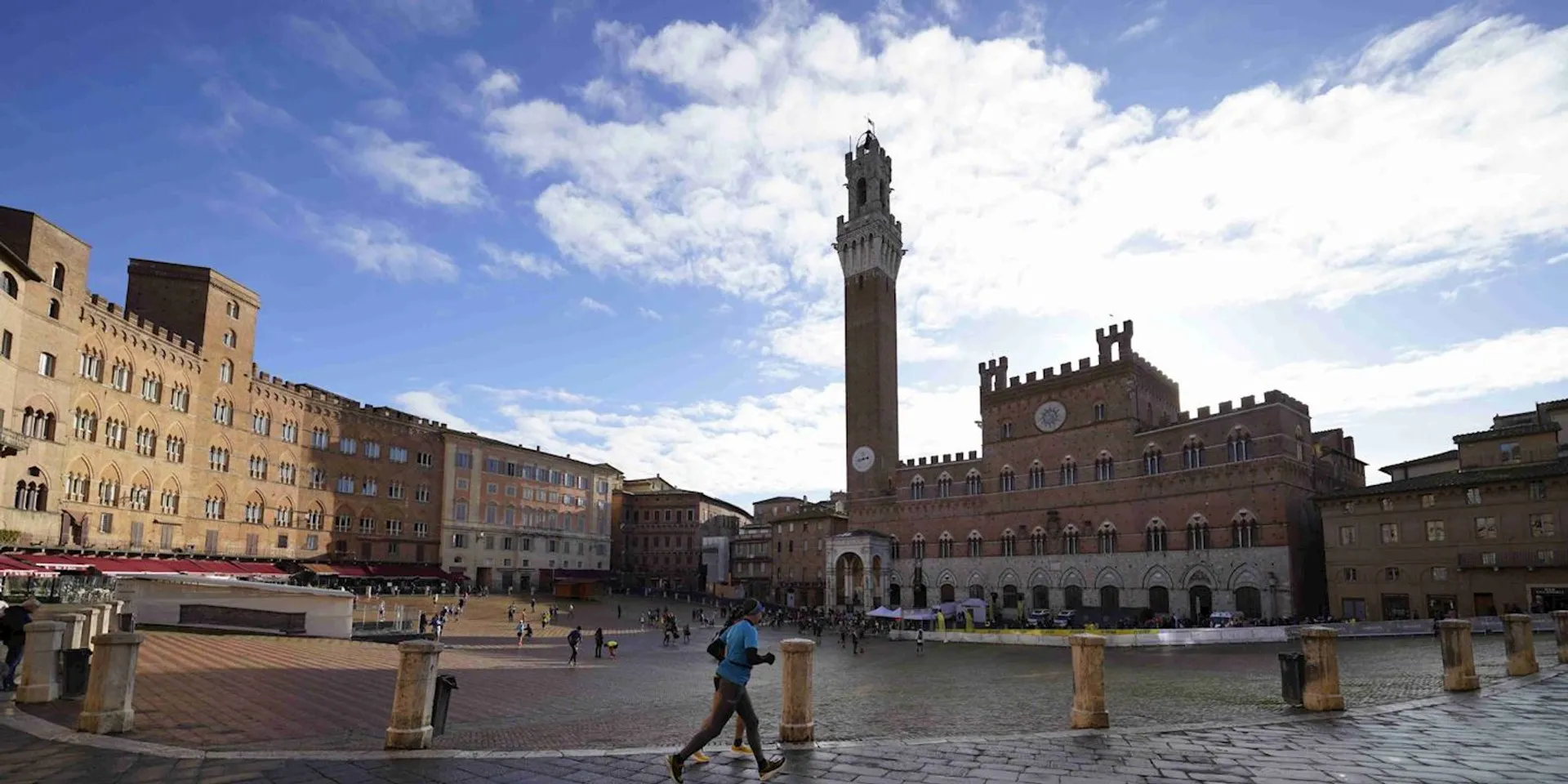 This image shows a large, open plaza with historic buildings surrounding it. Prominent in the center of the image is a tall tower, which is likely a bell tower or part of a town hall, given the architectural style. The buildings have a uniform, aged look, featuring what appears to be medieval or Renaissance architectural styles, typical of many European cities. The sky is mostly clear with a few scattered clouds, suggesting a pleasant day.

The ground is paved, and there are several people dispersed across the plaza, some at a distance and one closer to the foreground in motion, possibly walking or running. This could be a popular tourist location or a central square in a city, where locals and tourists congregate. The strong shadows indicate that the photo was