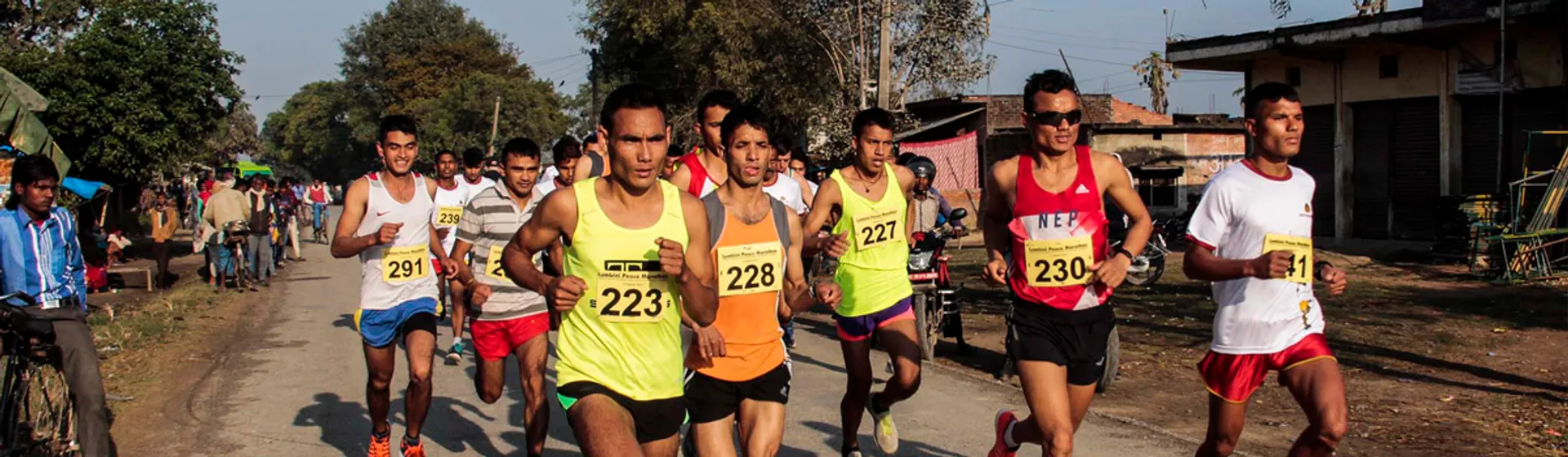 The image shows a group of male runners in a road race. They are wearing competition numbers pinned to their shirts, which suggests this is an organized event such as a marathon or a shorter distance race. Some of the runners are looking straight ahead with focused expressions, indicating that they are competing seriously. Spectators can be seen on the side of the road, watching the event. The setting appears to be in a place with warm weather, given the clothing of both the participants and the spectators, and there are trees and clear skies in the background.
