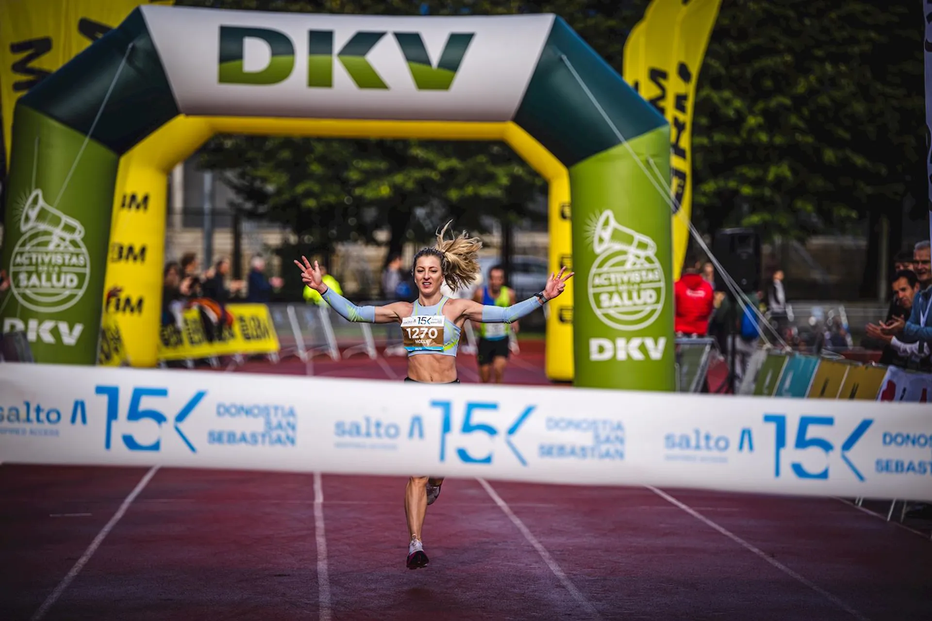 The image shows a female runner crossing the finish line at a race event. She is raising her arms in celebration. The event seems to be a 15K race held in Donostia / San Sebastian, as indicated by the signage at the finish line. There are also banners and people in the background.